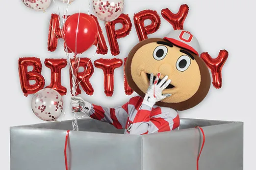 Brutus Buckeye puts a hand to his mouth as he giggles while rising from inside a giant gift-wrapped box.