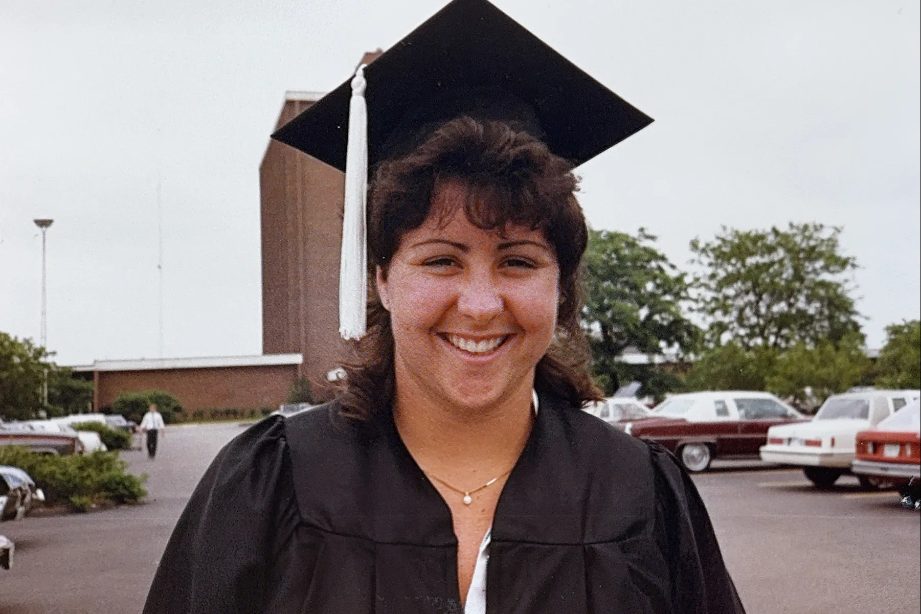 Molly smiles wearing her college graduation cap and gown. She's in the parking lot of Longaberger Alumni House and the tower of Fawcett Center can be seen in the background.