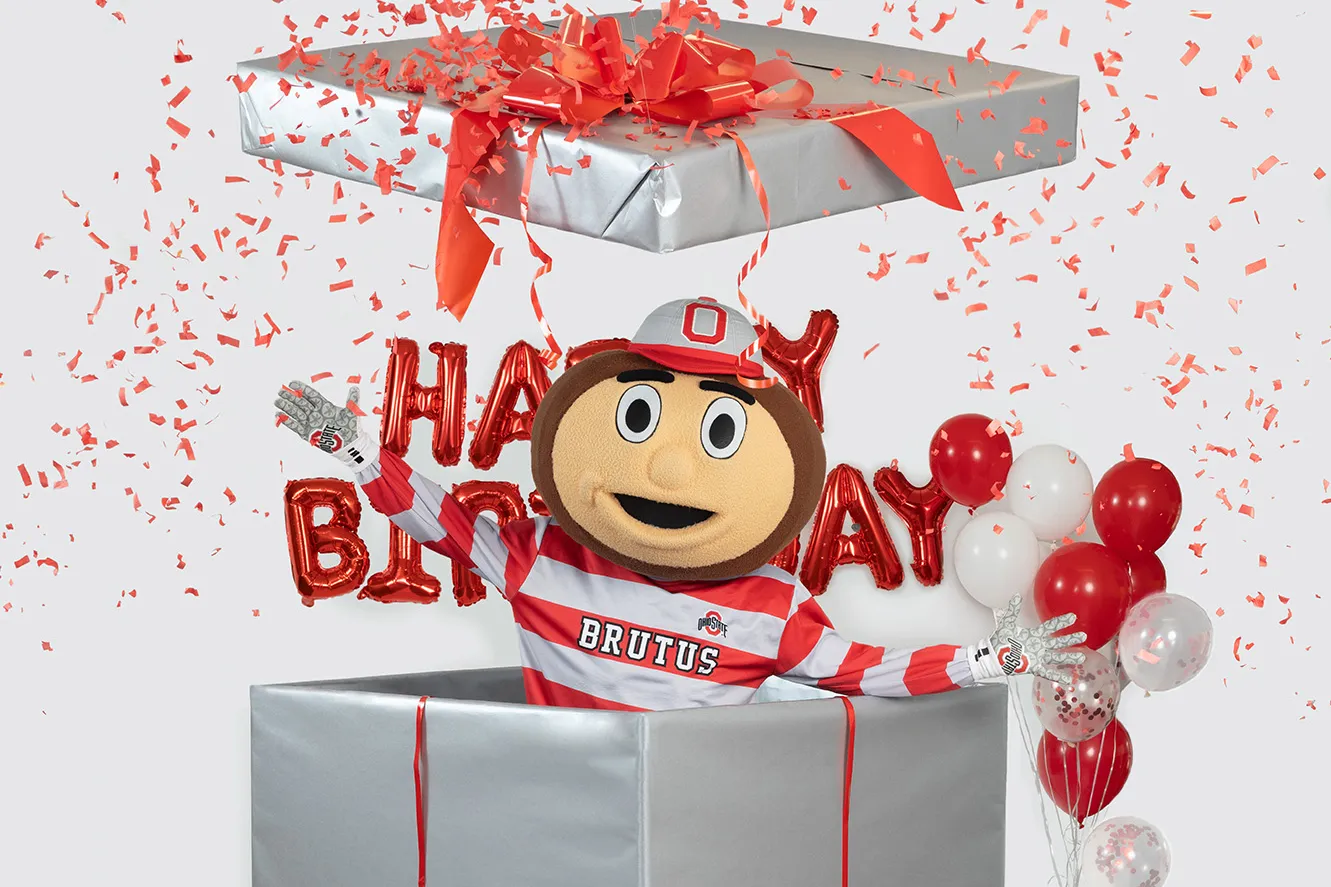 In this Ohio State photo, Brutus Buckeye jumps out of a giant wrapped birthday present. Balloons in Ohio State's colors, as well as shiny mylar balloons that say happy birthday, are around him and confetti flies everywhere. Brutus is the university's mascot--a giant head shaped like a buckeye tree nut with a big grin.
