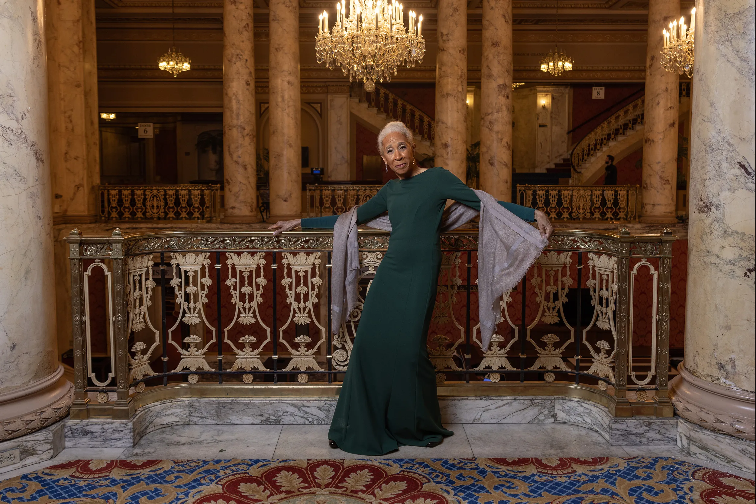 Dianne McIntyre, a slender and stylish black woman, poses along an ornate railing in a historical theatre with big marble columns and intricate detailing. Dianne is an attractive older woman. She has white hair pulled back in a bun, long legs and her body is curved as if in motion, with her arms outstretched. She wears a sleek column dress and one hand holds the end of a silk wrap draped around her back. 