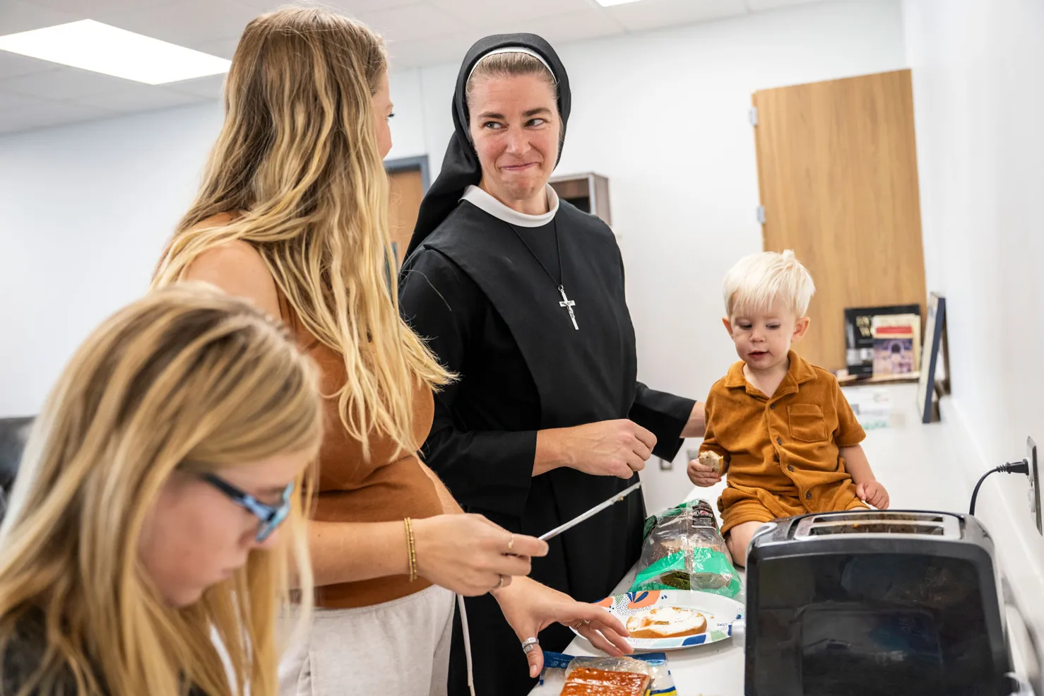 Sister close-mouthed smiles at her biological sister as they make sandwiches at a counter. A young boy sits on the counter; sister supports him with one hand. And older girl stands next to her mom. They all have blond hair and seem to be enjoying each other's presence.