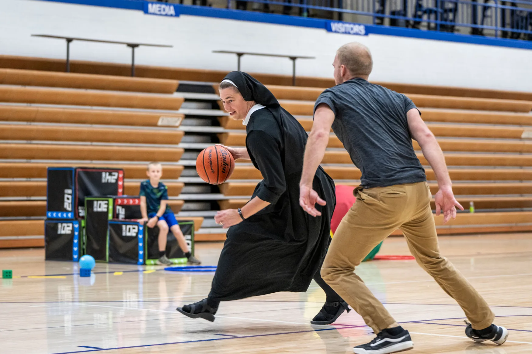 Sister dribbles as her brother-in-law guards her on a basketball court. A boy watches from the sideline.