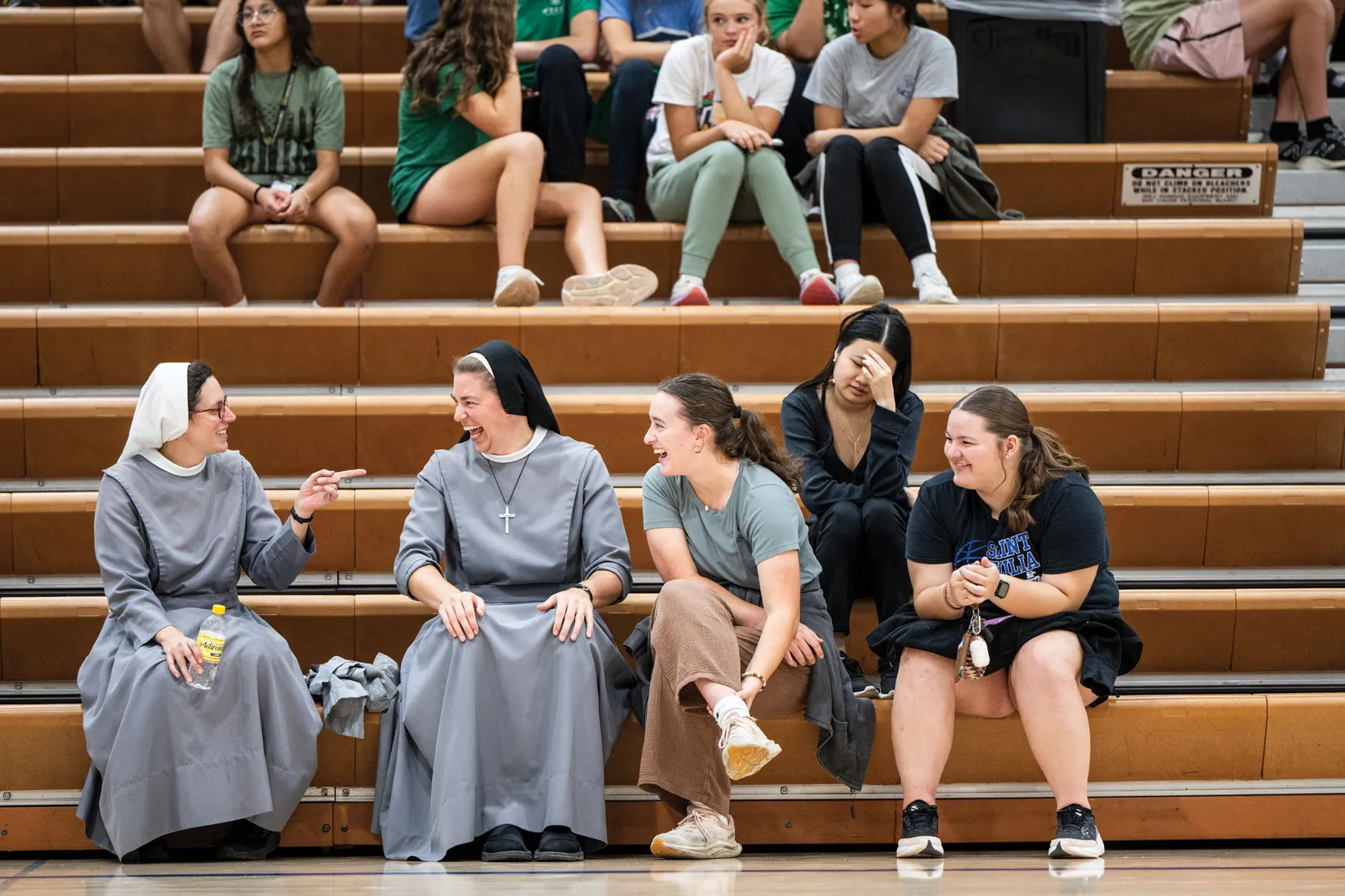Two sisters sit on bleachers in a gymnasium laughing as teenagers sit around them.