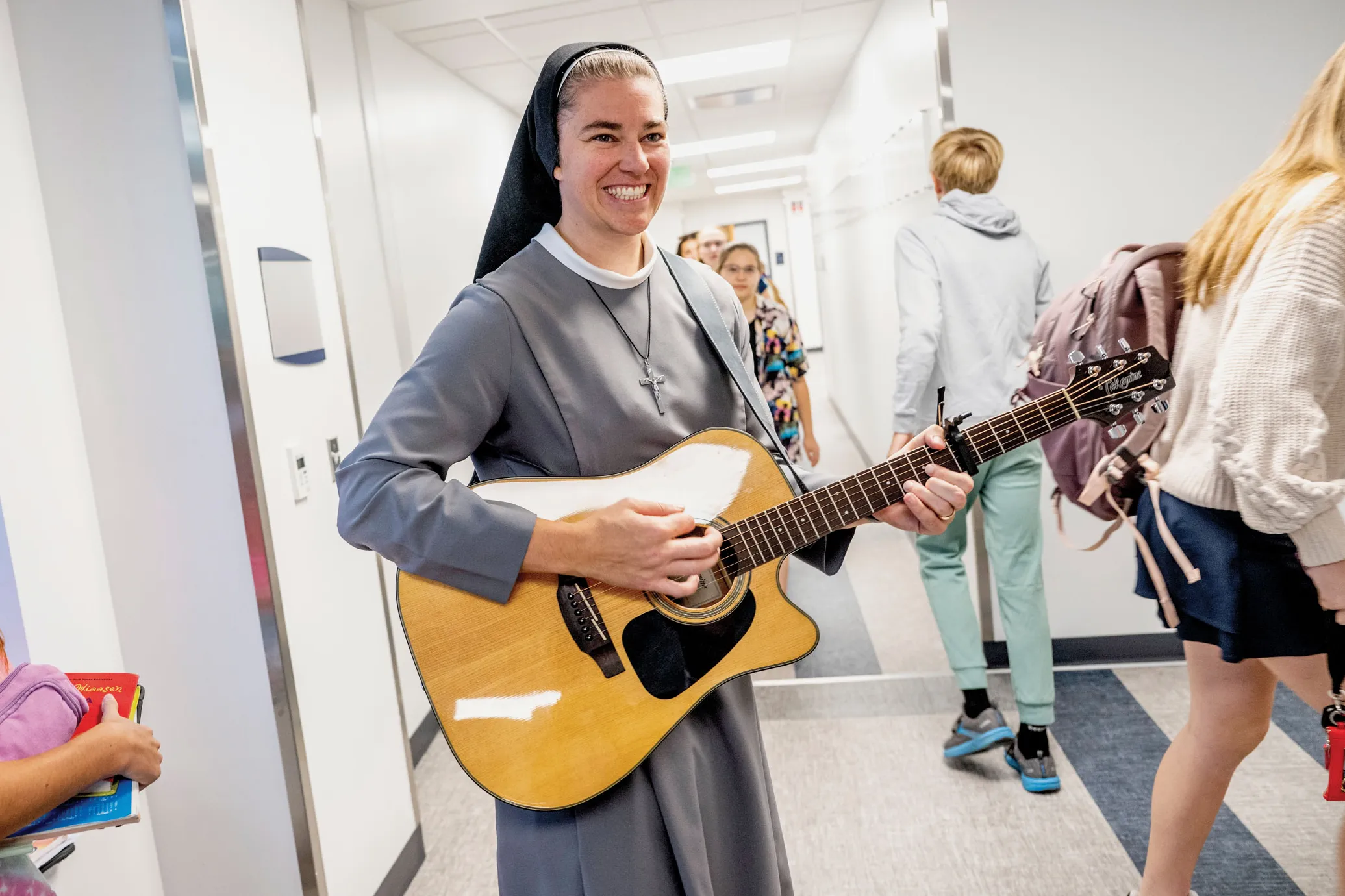 Sister Mary Xavier Schulze is a white woman wearing a veil and habit in shades of gray, white and black. She smiles big and plays a guitar as she walks the halls of the school she works in, as kids rush past her.