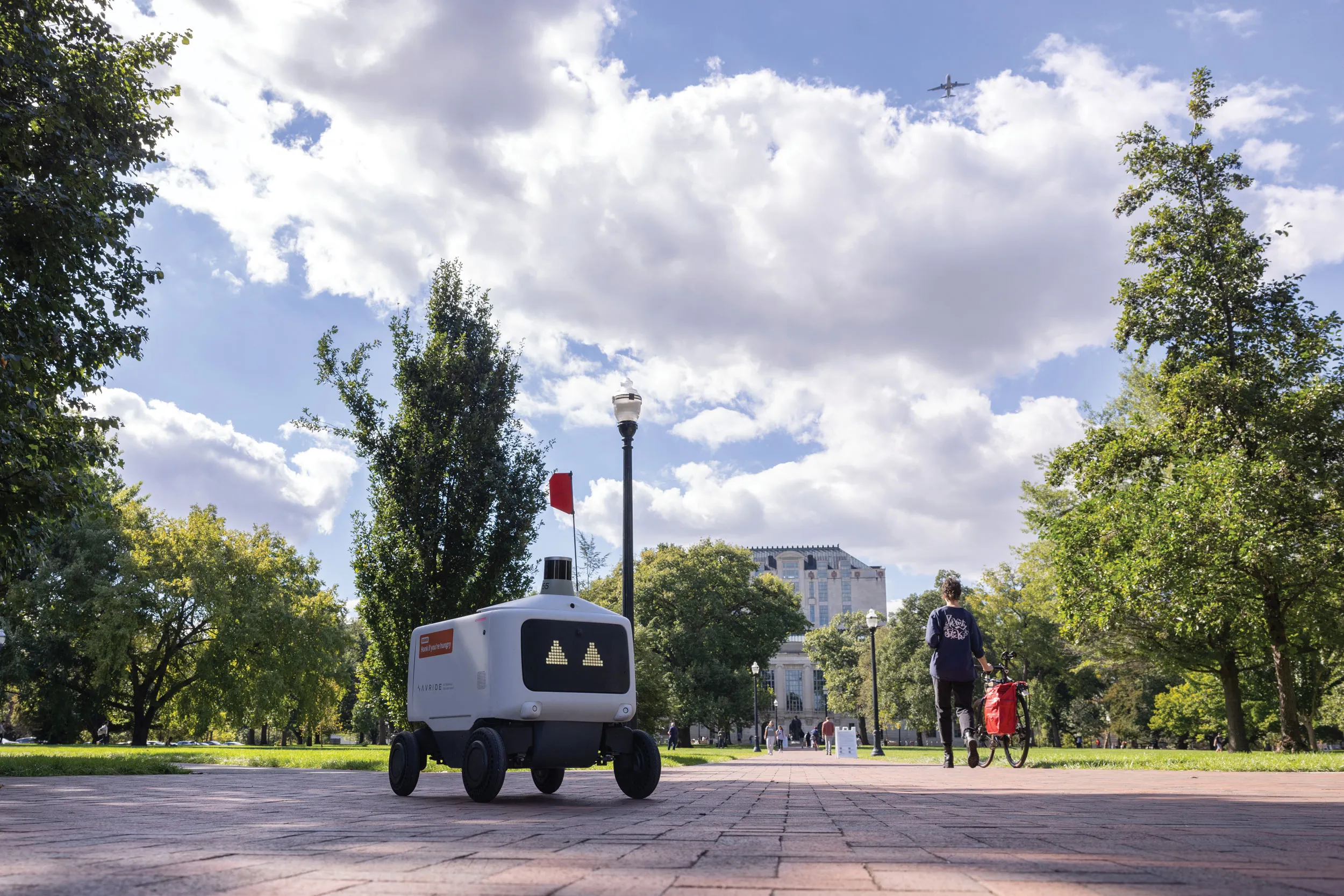 A lone food rover, which is a plastic-covered rectangle with a screen face showing triangle-shape eyes, makes its way across the oval. Thompson Library and students can be seen in the distance.