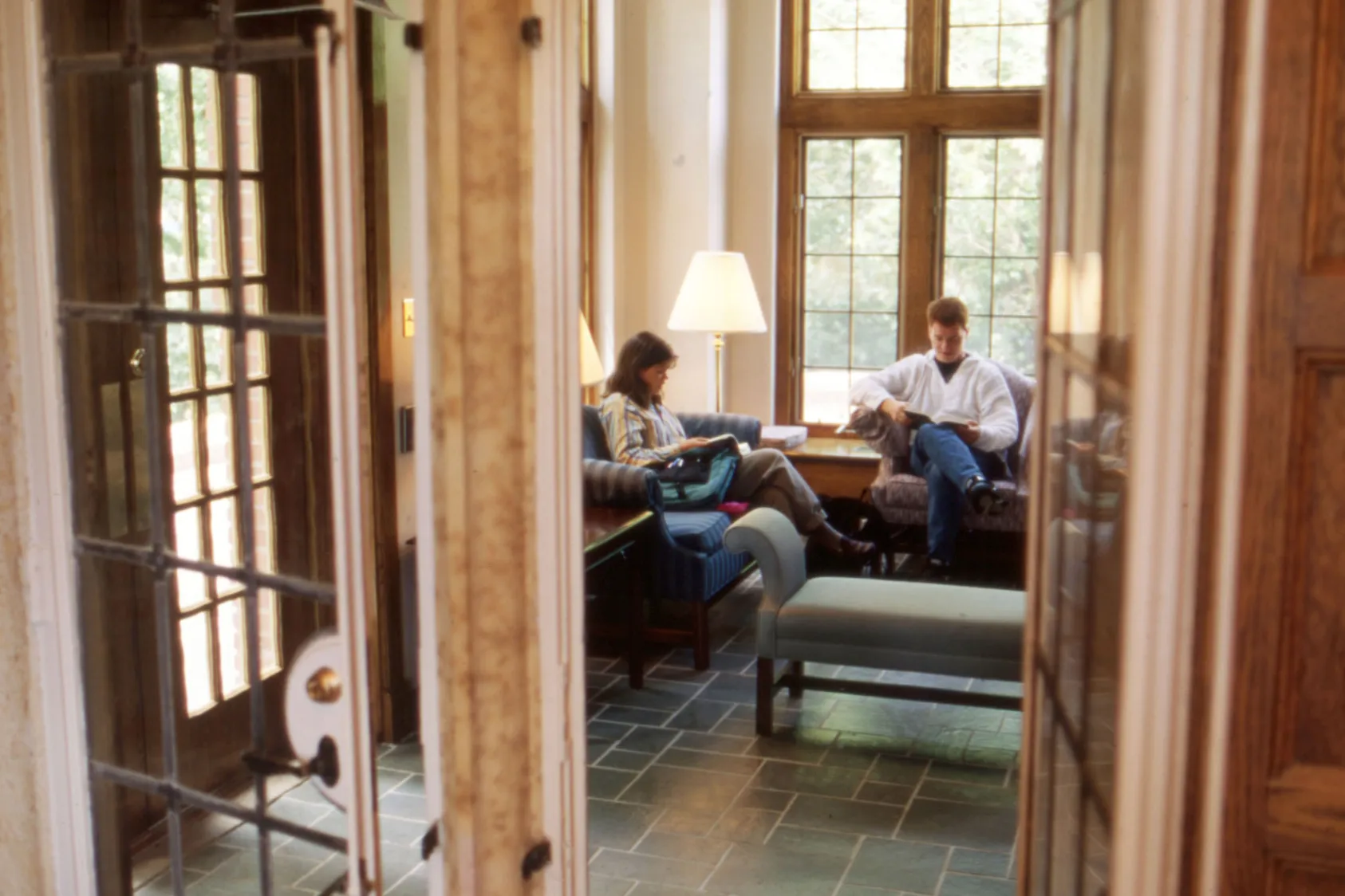 Two people sit in a lounge area near large windows. They face each other while seated on cushioned furniture, with notebooks or papers in their laps. The scene is viewed through open interior doors, showing tiled flooring and wood-paneled walls.