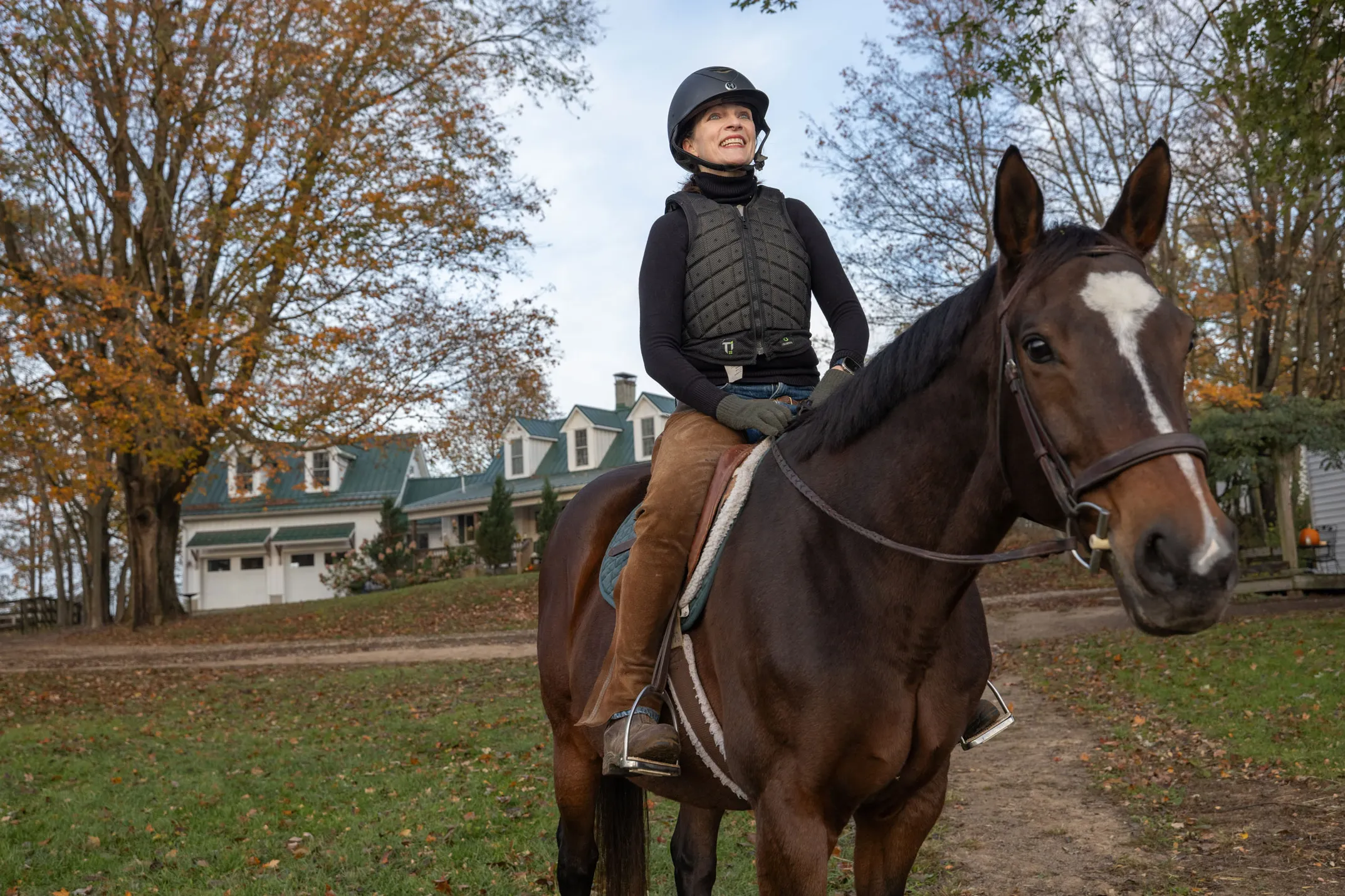 Kathy Lloyd smiles as she sits on a horse outside her riding stables on a crisp autumn day. She wears a helmet and protective riding vest sits and the horse looks directly at the camera as she looks off into the distance.