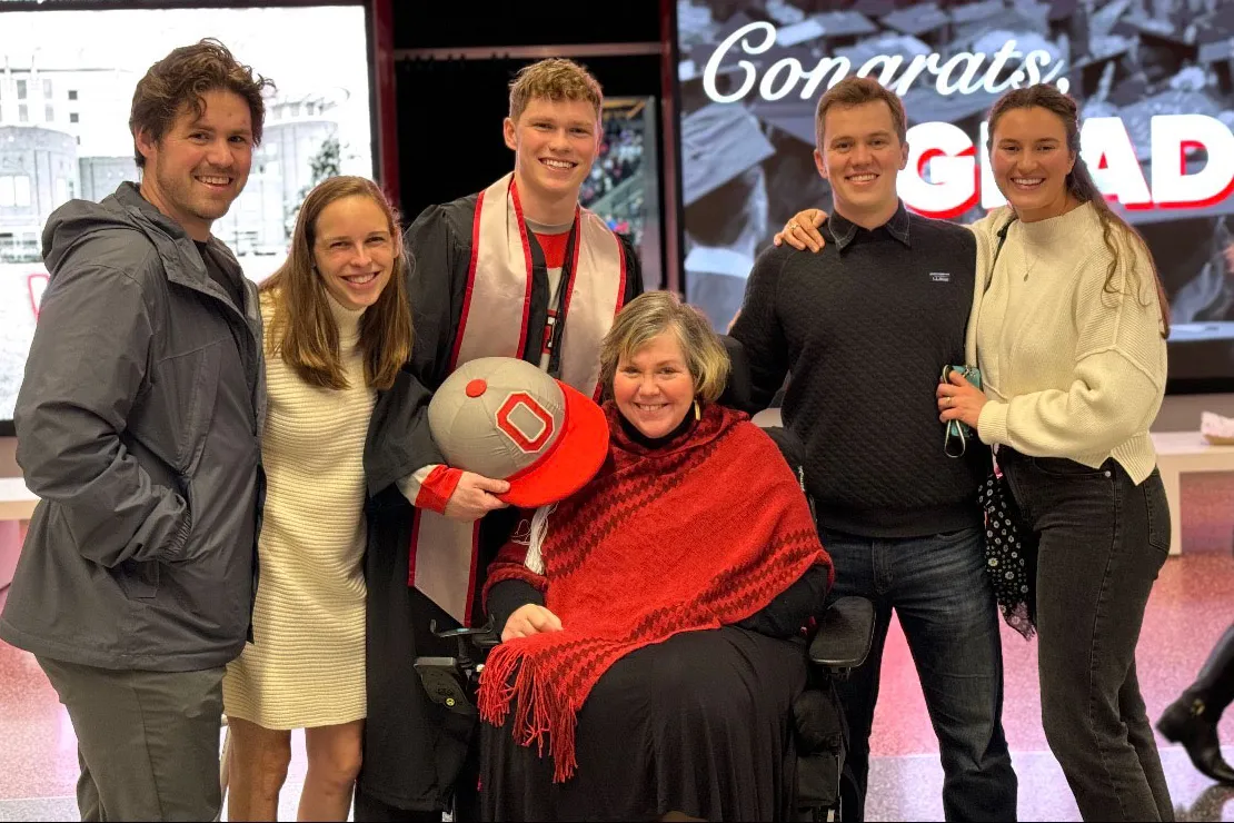 A family gathers around a mom sitting in a wheelchair. One of the 5 people standing behind her wears a graduation gown and holds his graduation cap, which is topped by Brutus Buckeye's giant cap.