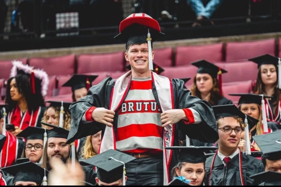Graduates sit in stands inside Ohio Stadium. One young man stands up, separating his graduation robe to show off his striped Brutus shirt. On his head, his graduation cap is topped by Brutus' hat. His grin is proud.