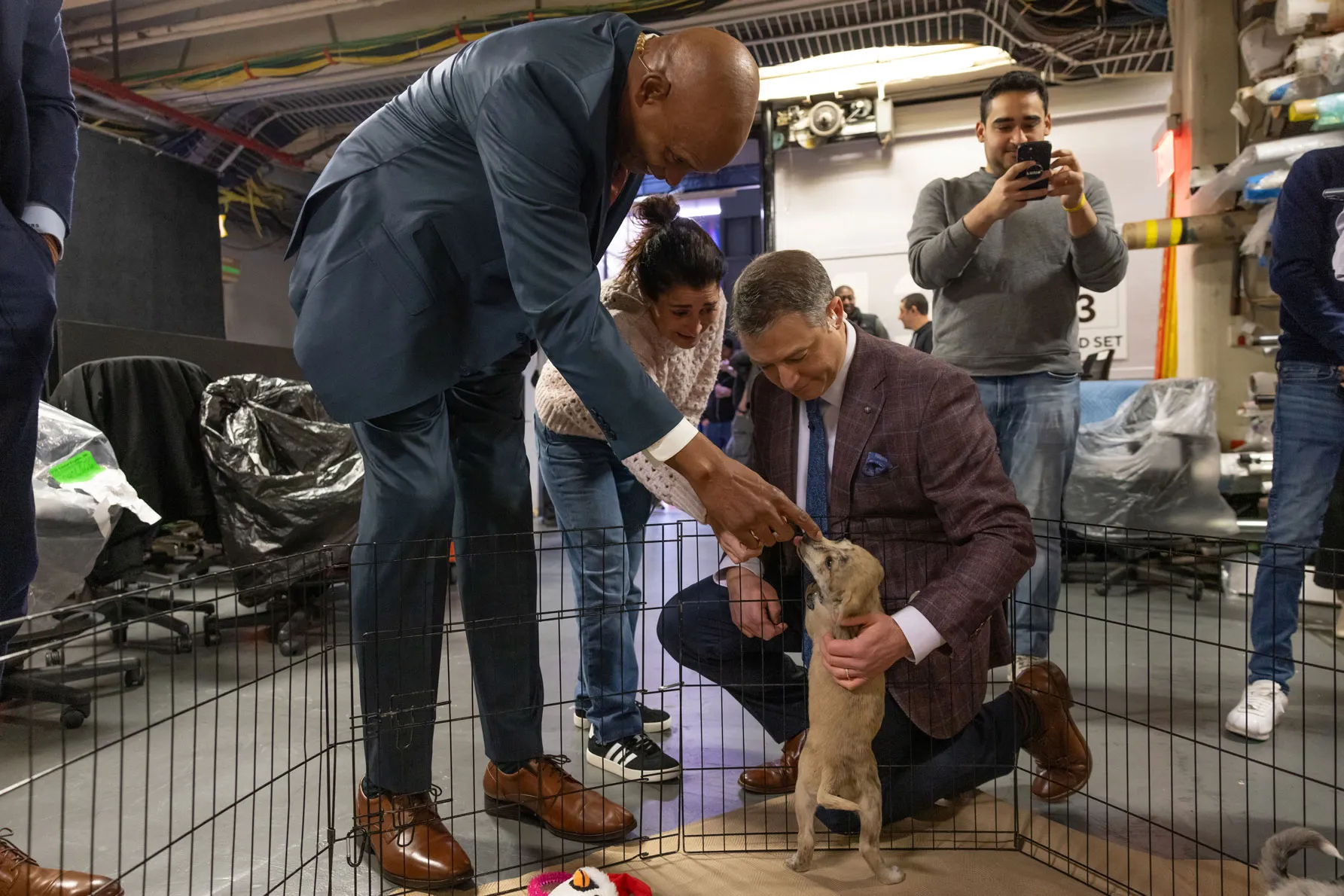 Kellogg, Zucker and more studio workers gather around a small fenced play area in a studio workspace, interacting with a small dog standing on hind legs. The incredibly tall Kellogg leans over to let the puppy sniff his hand.