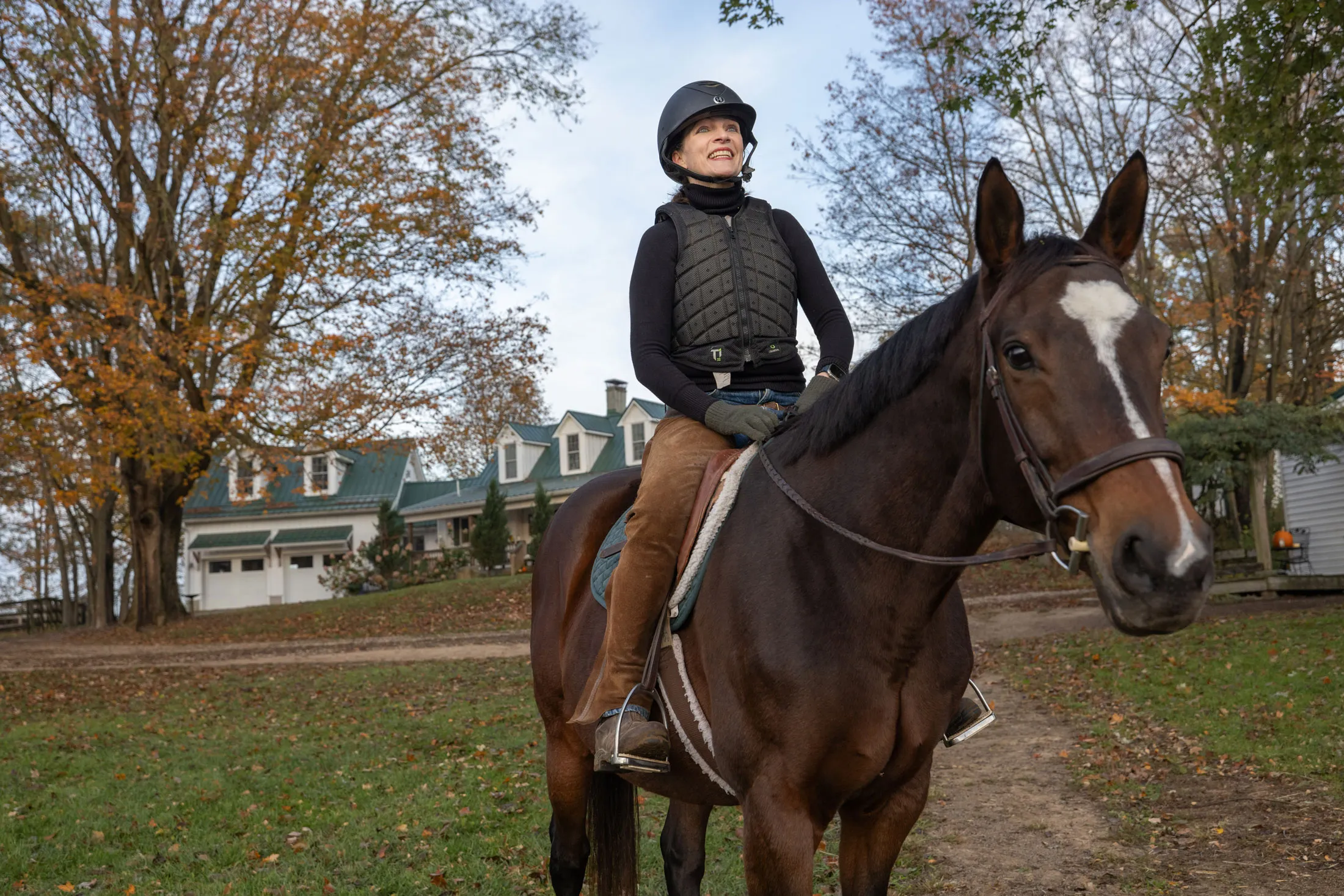 Kathy Lloyd smiles as she sits on a horse outside her riding stables on a crisp autumn day. She wears a helmet and protective riding vest sits and the horse looks directly at the camera as she looks off into the distance.