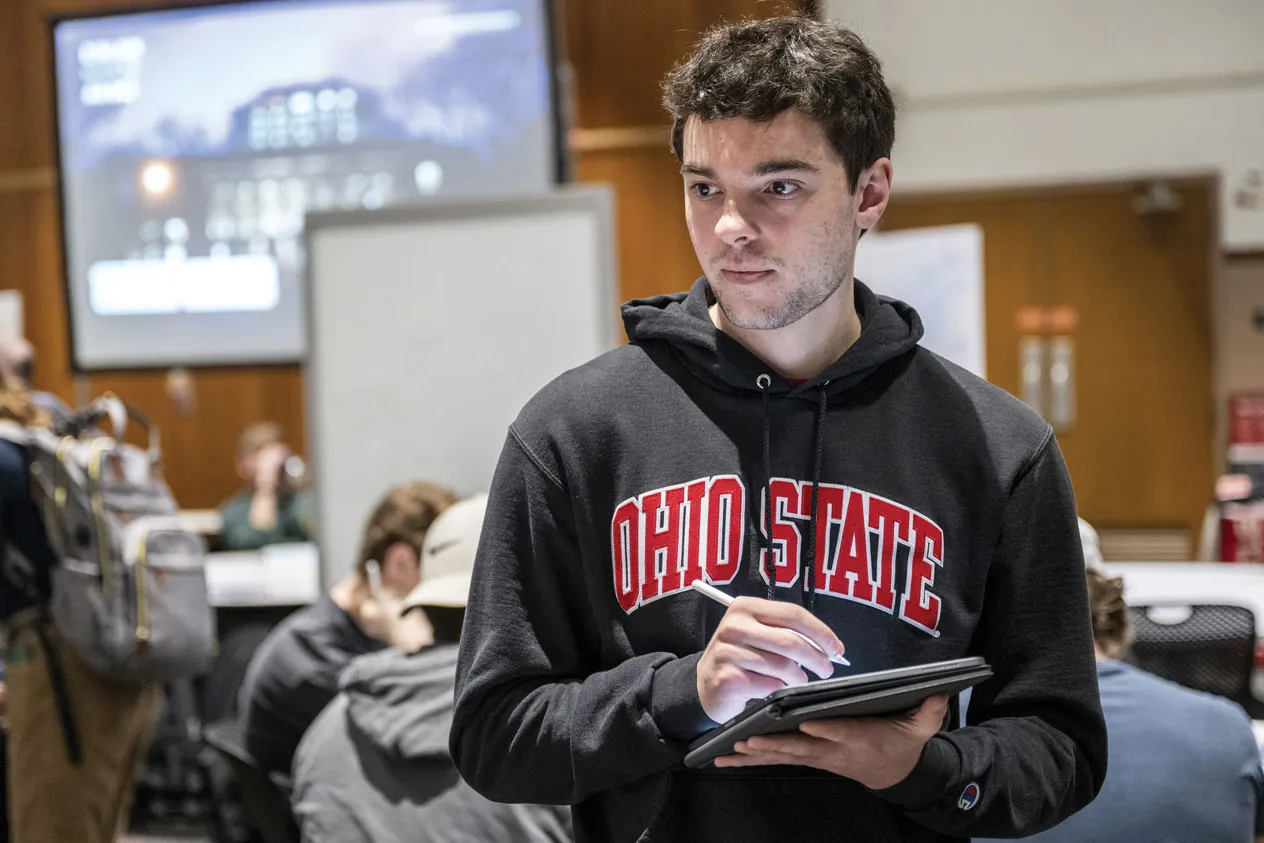 A student takes notes as classmates talk about his work. He wears an Ohio State sweatshirt and an intensely focused expression