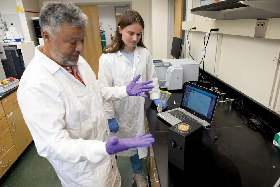 Professor Luis Rodriguez-Saona gestures with hands wide and fingers spread, as he and PhD student Fiona Schmidt watch the spectrometer work.