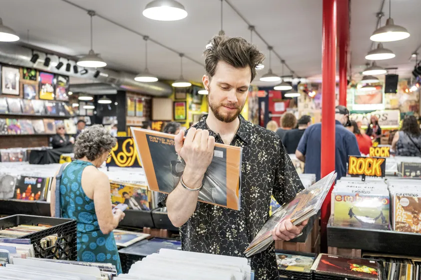 A patron with cool, messy hair considers the back of an album cover, while holding several more records in his other hand, as if those are the ones he has decided to purchase
