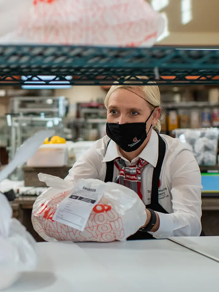 Abby Hertzfeld is holding a take-out food order wrapped in a white plastic bag. She is wearing a black face mask and hairnet.