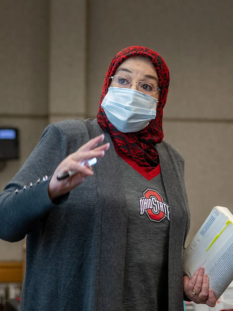 Rachida Labbas is standing with a book in one hand and a marker in the other. She is wearing a gray OSU shirt and cardigan, a red and black hijab, a face mask and glasses.