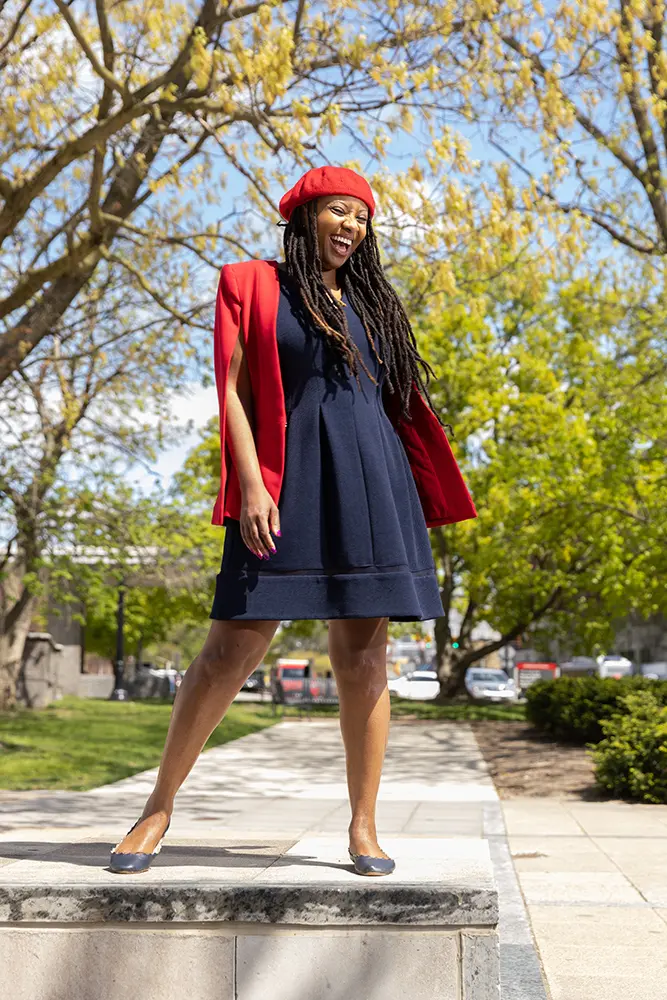 A woman wearing a dress and a beret laughs while outside on a pretty day. 