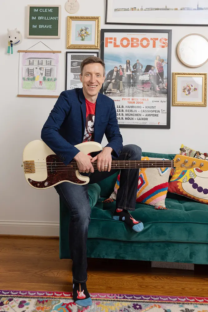 A man whose outfit includes a suitcoat and guitar-patterned socks holds his base guitar and smiles while perched on the arm of a couch.