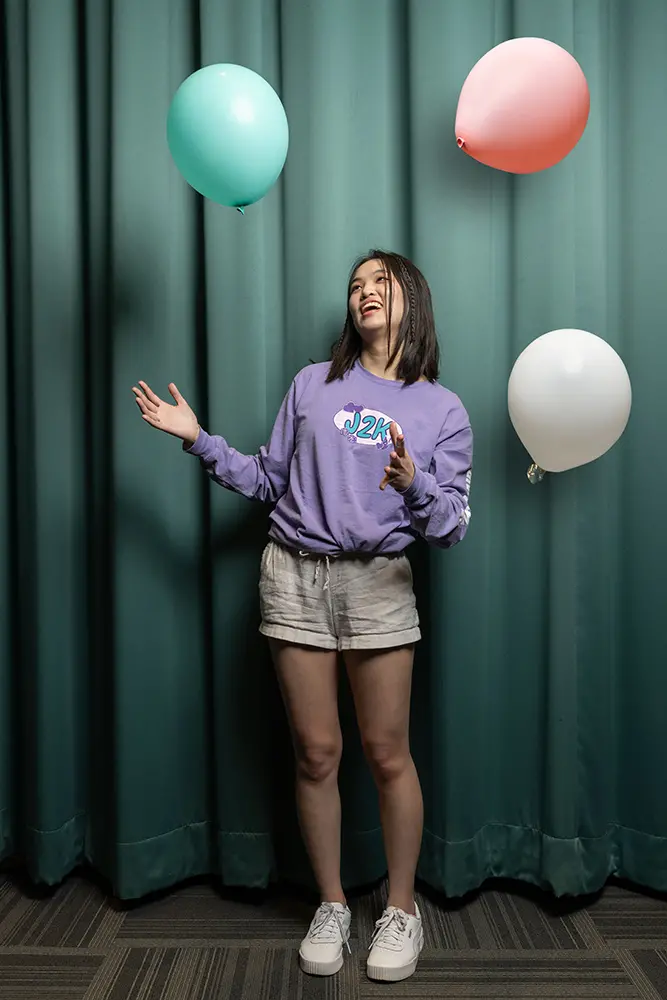 A young woman stands in front of a stage curtain with balloons in the air around her. She wears a J2K T-shirt and shorts.