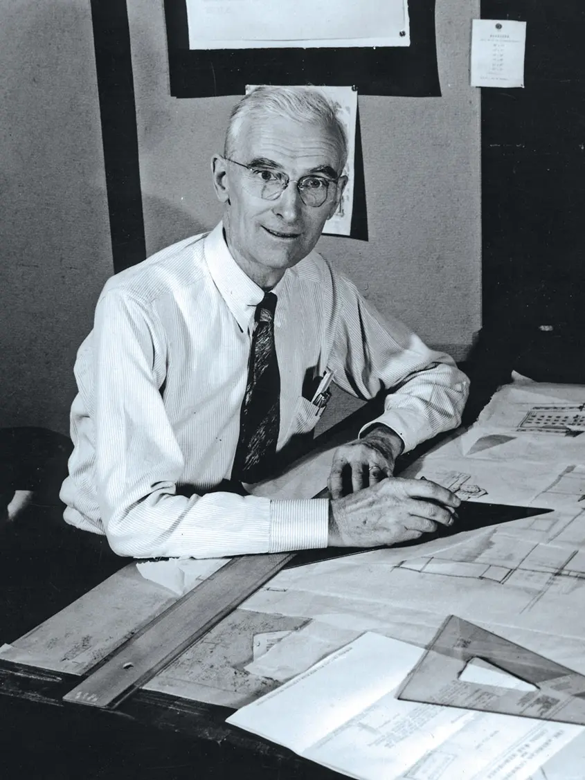 A man with white hair, weaing a button-down and tie, works at a drafting table on an architectural drawing. The photographer seems to have caught him off-guard but in a good mood.