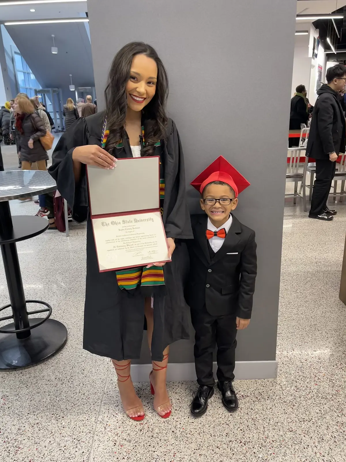 A young black woman in a graduation robe smiles brightly as she shows her diploma, and her son, who seems to be no older than 5 or 6, grins. He’s wearing a little suit with a scarlet bow tie and graduation cap.