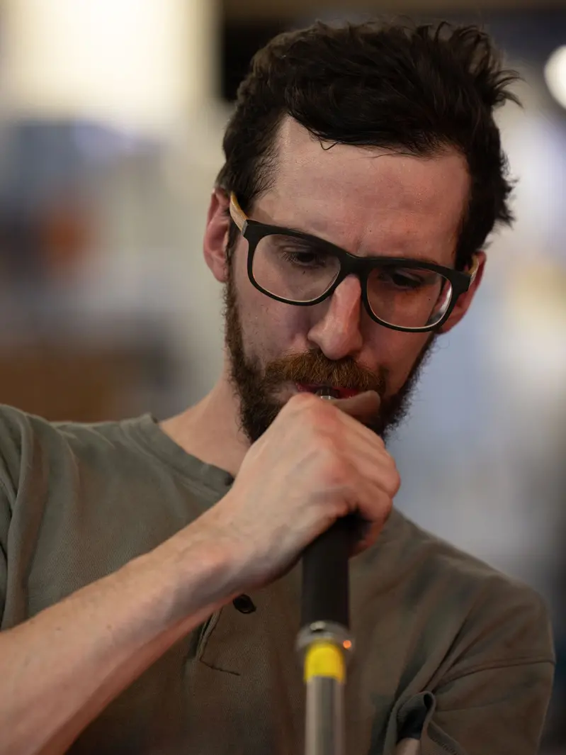 A white man wearing thick-rimmed glasses looks focused as he blows into a pipe used to inflate hot glass. The artist looks hot and tired — his short hair is messed up, as if he has run his hands through it many times.
