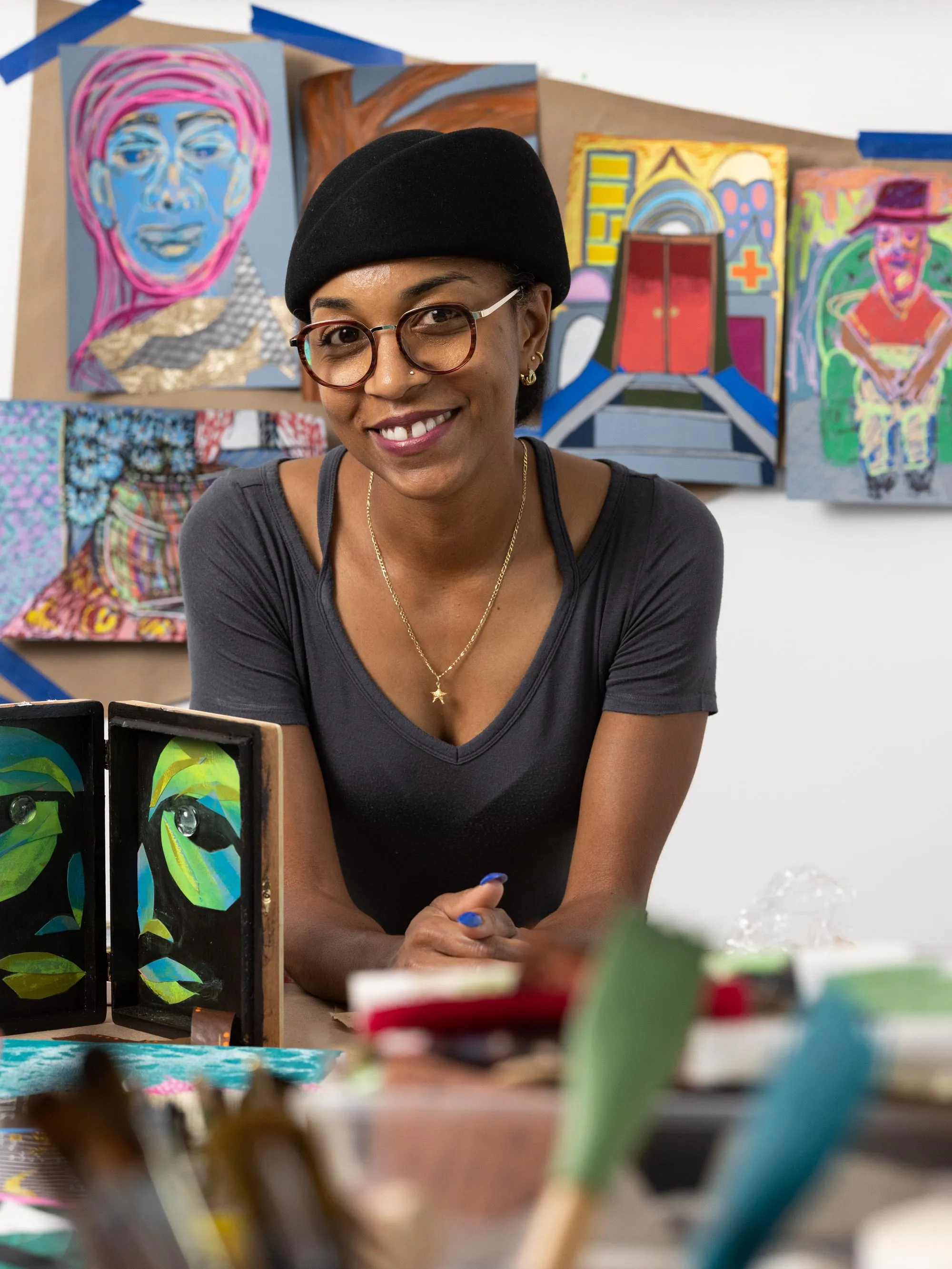 Tiffany Lawson, a Black woman wearing a hat, round glasses and a friendly smile, leans on her work table. In front of her are all kinds of supplies and works in progress and on the wall behind her are paintings she created on paper bags. They include people, faces and scenes in unexpected colors. 