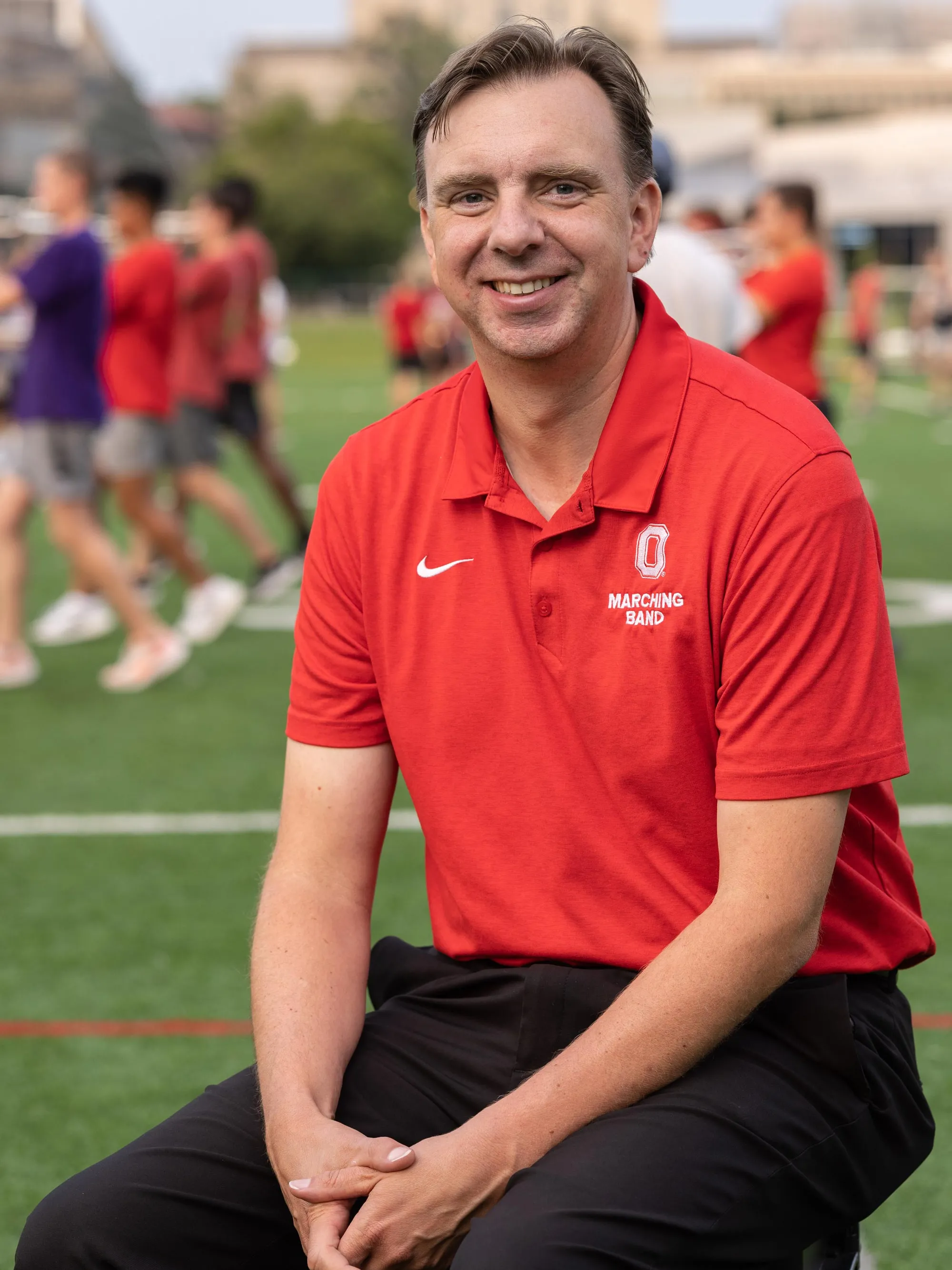 As members of the band practices behind him in their street clothes, marching band director Chris Hoch sits on a stool on the practice field. His hair’s a bit messy, his clothes are casual, and his expression is happy. 