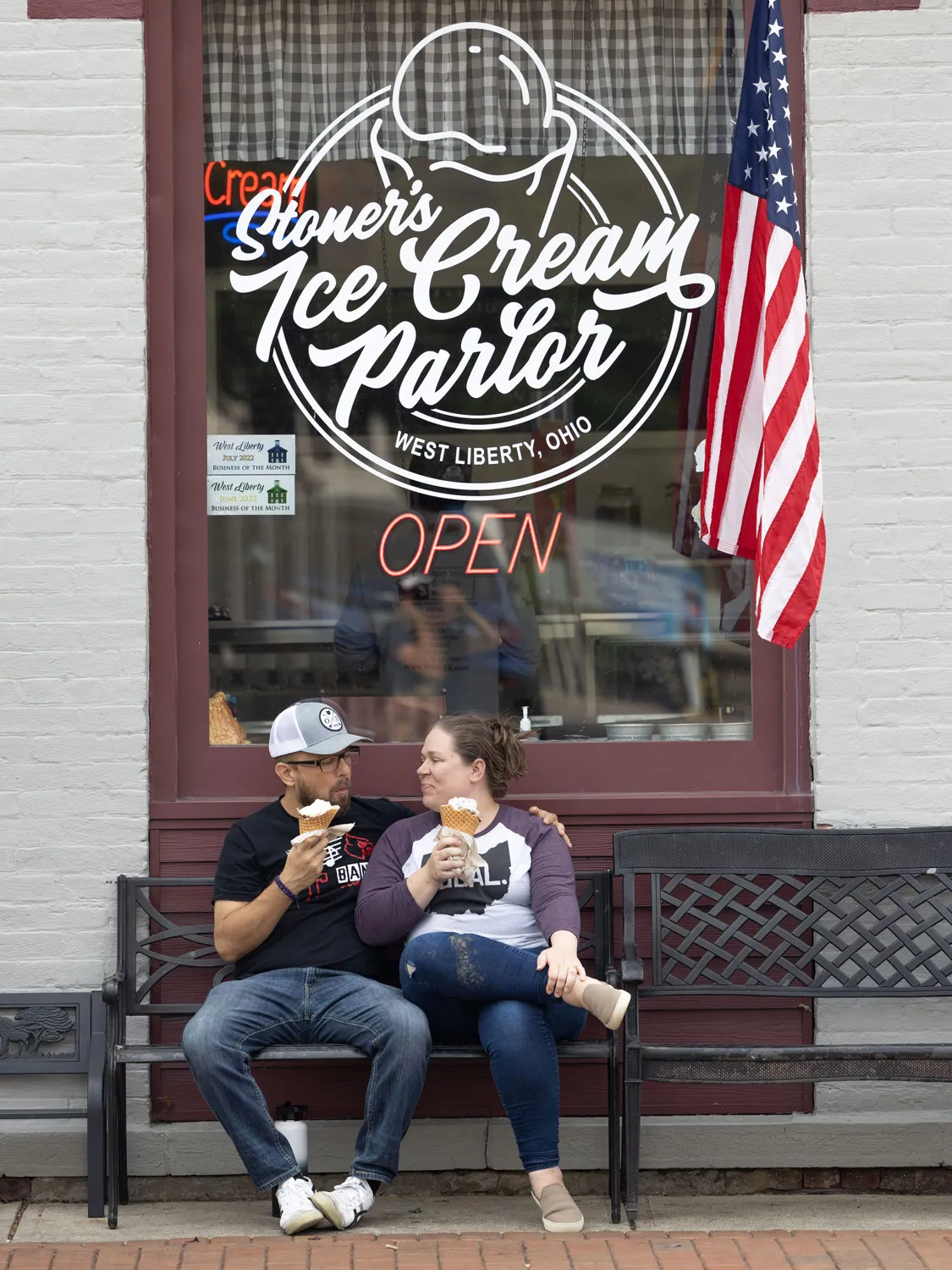 Gretchen Klingler sits with her husband on a bench in front of Stoner’s Ice Cream Parlor in West Liberty, Ohio. The name is painted in script on a huge window behind them, of the white-painted brick building. An American flag is mounted on the window frame. Gretchen and Aaron Gall, a white man with close-cropped beard and hair, both wear T-shirts and jeans and hold waffle cones. They’re looking at each other and smiling. 