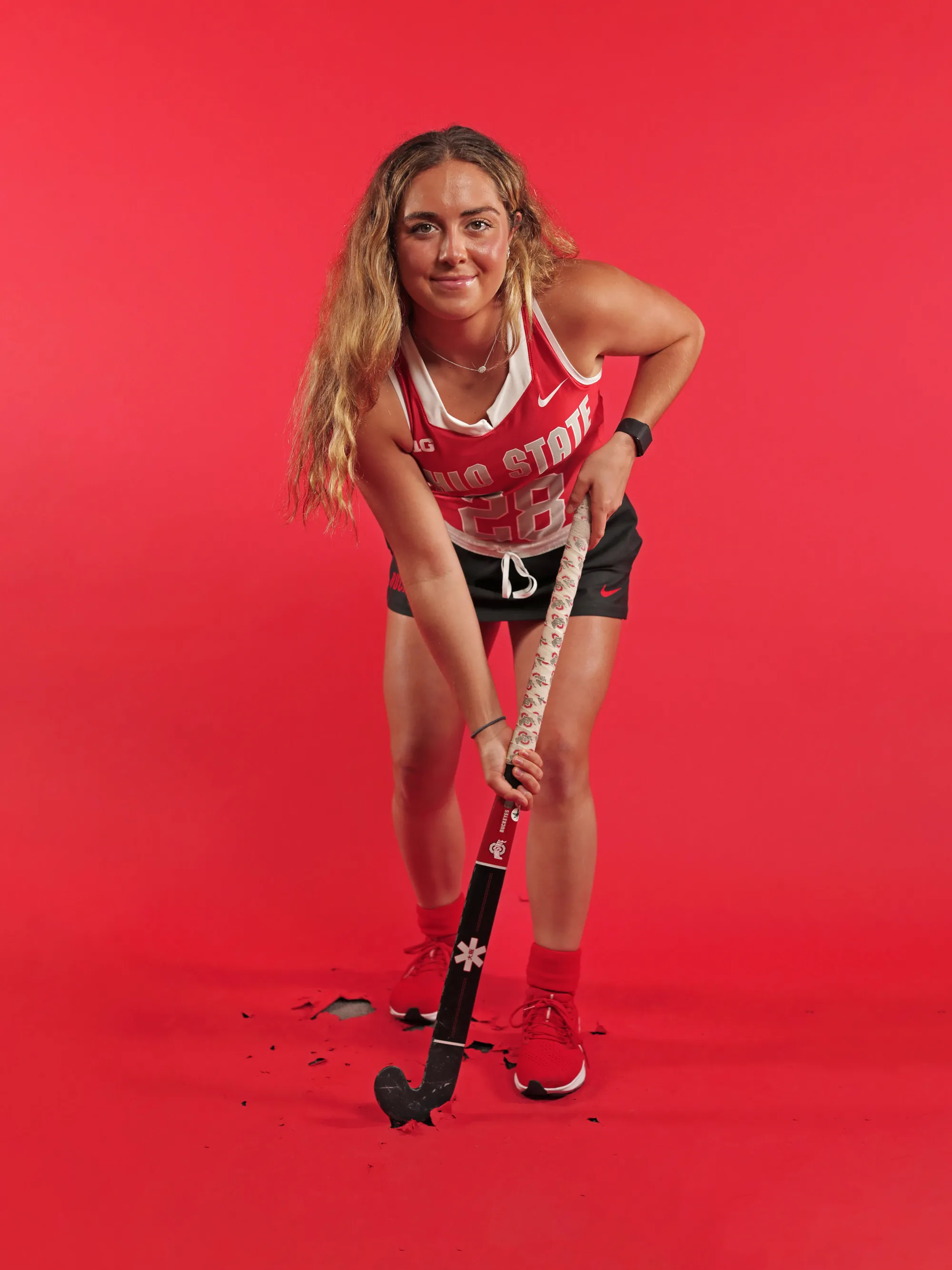 In her field hockey uniform with her stick, Makenna Webster bends forward in a play-like way, with a slight smile and her long hair hanging down. 