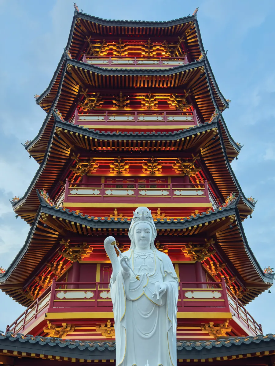 An ornate pagoda rises into the sky behind a smooth, white statue of a holy-seeming person on top of an elegant bird.