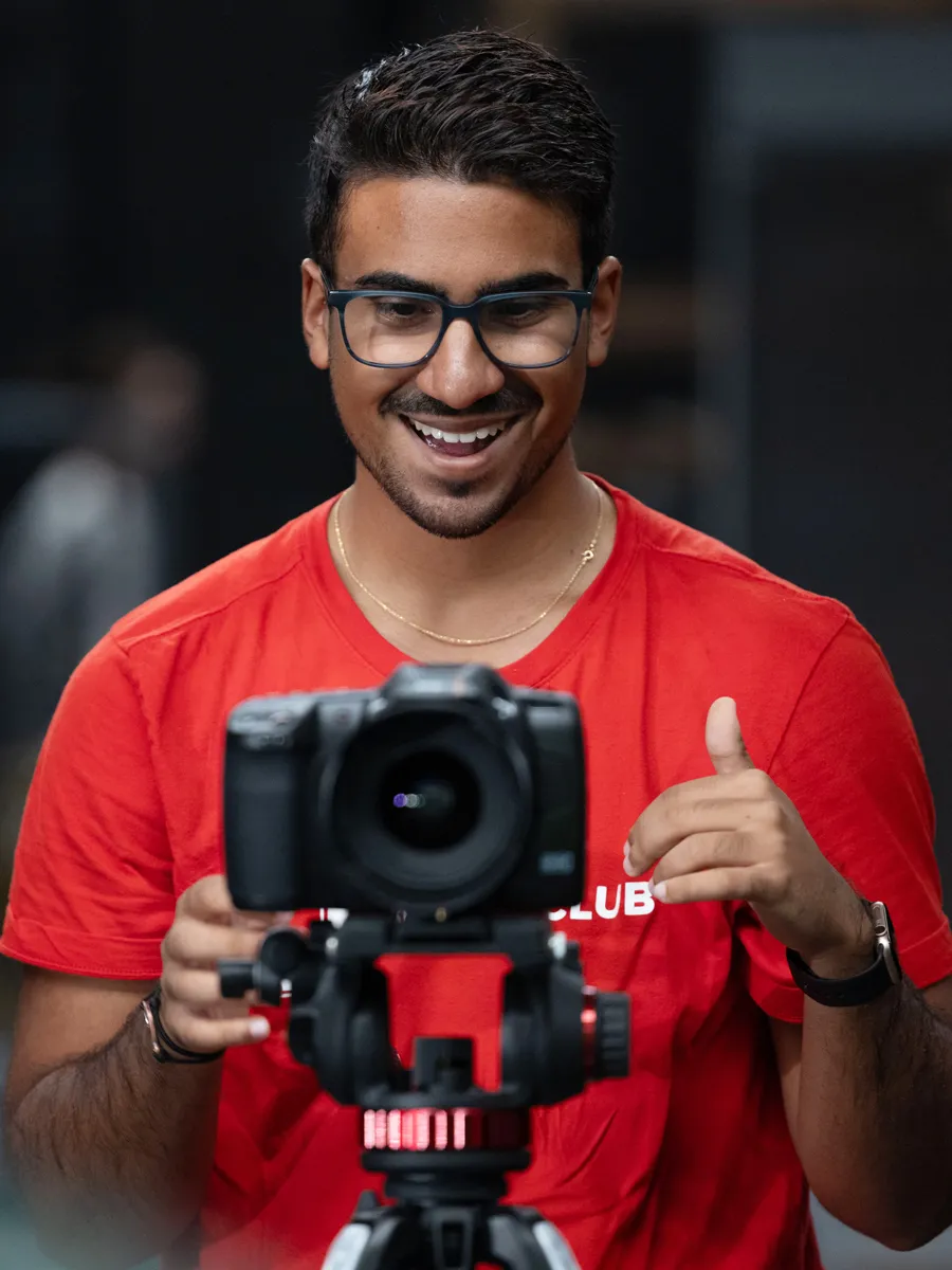 A young man laughs as he looks at the screen on the back of his camera, which is mounted on a tall tripod.