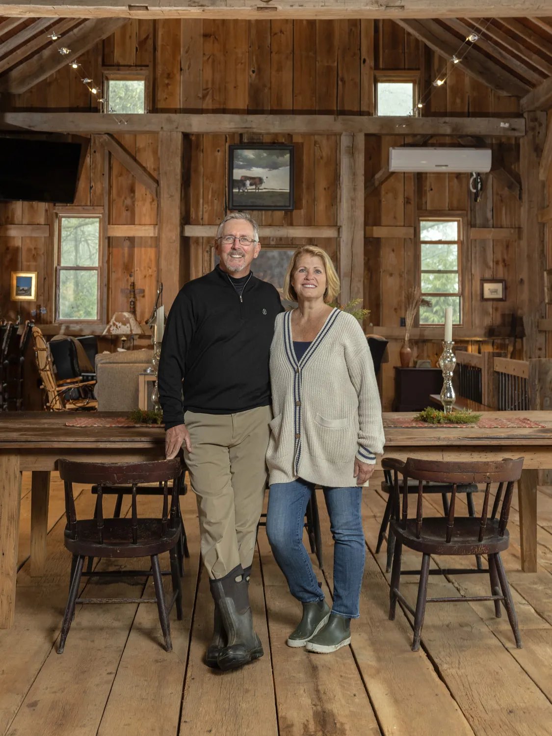 Standing inside a restored barn, a middle-aged couple wrap their arms around each other and smile for the camera. The structure, which peaks high above their heads, is made completely of wide planks of seemingly high quality lumber. There are paintings on the walls, windows and a couple of fancy, tall candle sticks and holders, giving an impression of upscale country. 