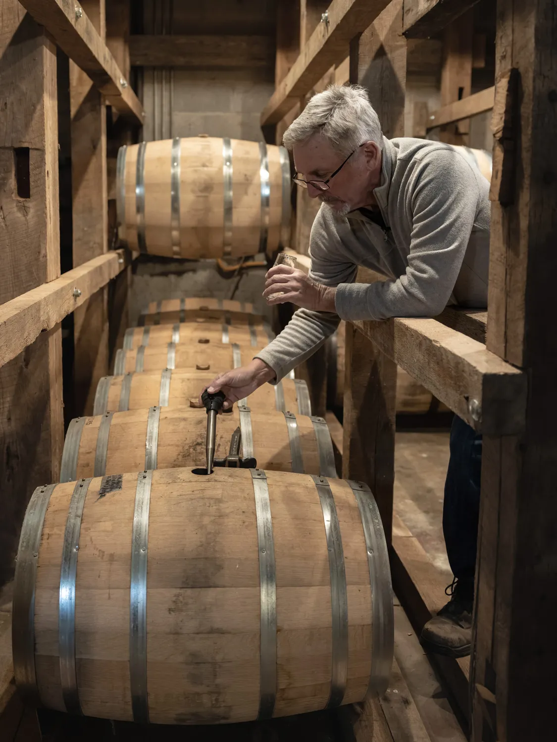Doug Morgan leans over a beam into an area where a line of whisky barrels lie on their sides. In one hand he holds a small glass cup; in the other, a pipette sized like a turkey baster to draw a sample of the alcohol. 