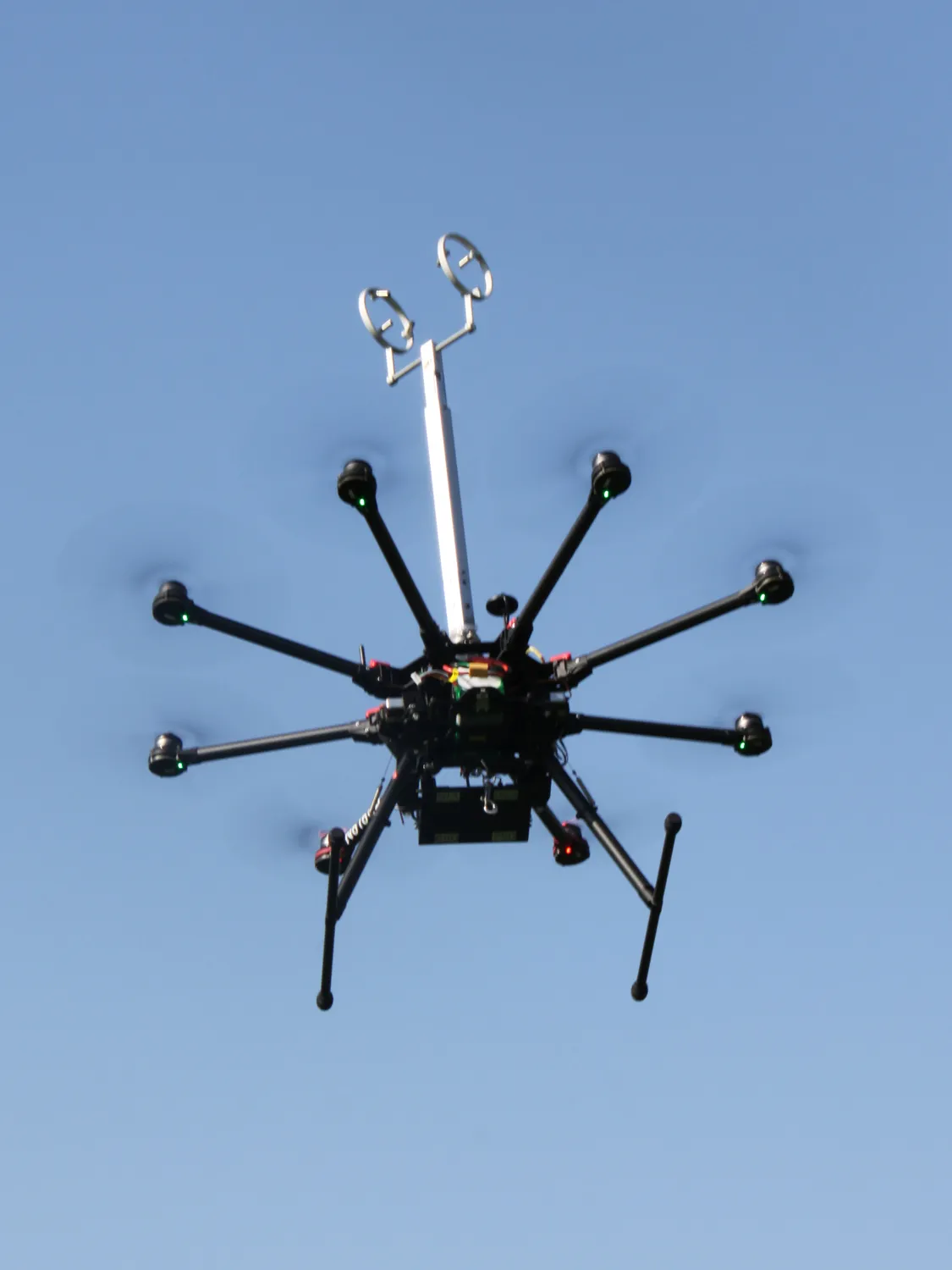 High in the sky, an eight-propellered drone can be seen from underneath. A metal antenna-seeming piece extends from the tops, at least four times taller than the drone’s body. 