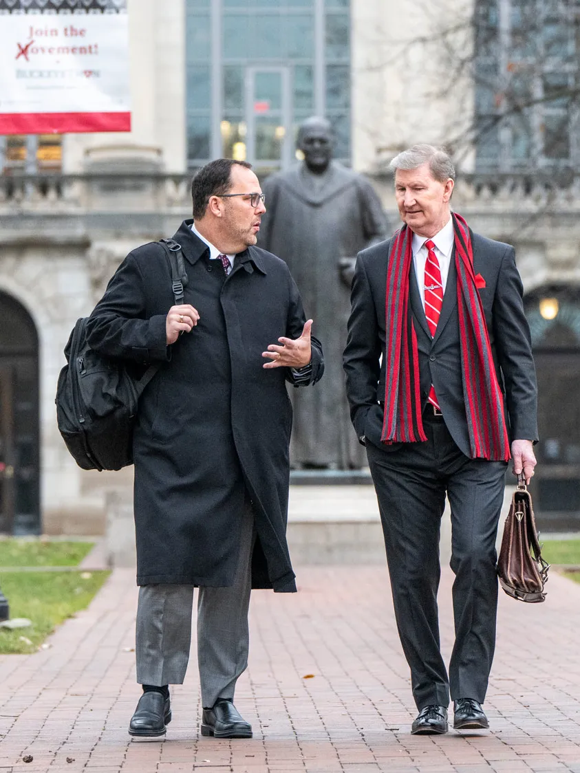 Ohio State President Ted Carter crosses the Oval with a confident but slight smile as we walks next to a university official. Carter wears a suit and long staff striped in Ohio State colors. JR Blackburn wears a black work coat and carries a backpack. In the background are stately academic buildings. The trees are bare but the grass is green 