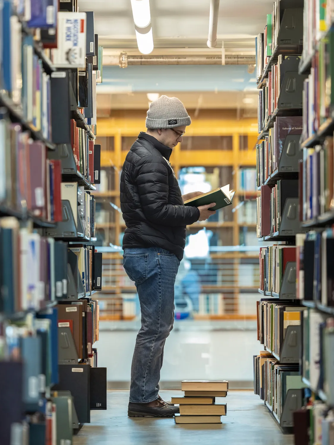 A white man in winter coat and hat focuses as he pages through a book in the stacks (which means he is standing between two closely placed rows of shelves, each chock full of research books). He is near the glass wall that looks into the main lobby. Five thick books sit in a stack at his feet. 