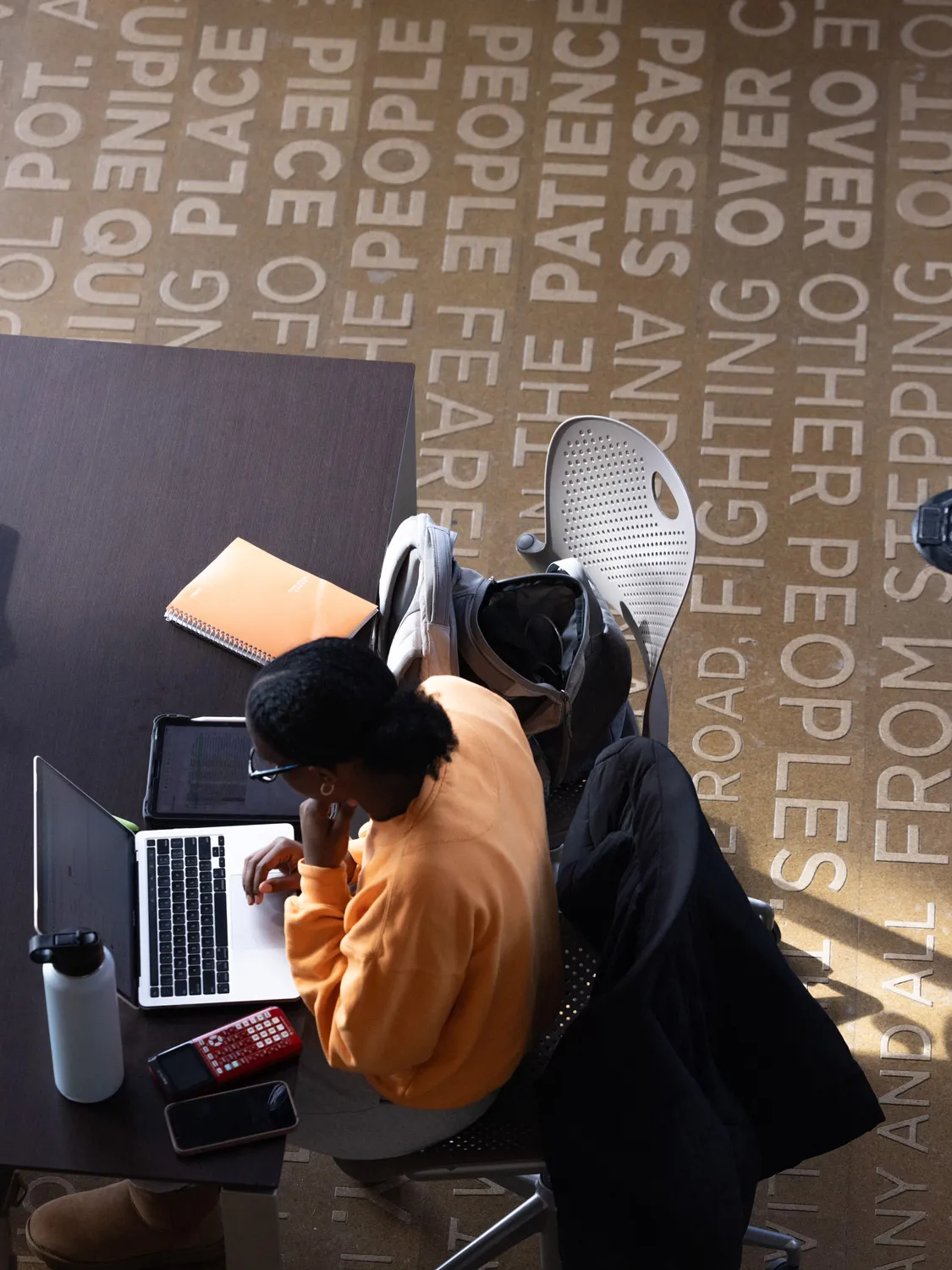 A young Black woman sits at a table working on her laptop. She is photographed from above, and the color of her sweatshirt matches her notebook. Just as prominent in the photo is the floor, which has words in lines, some that the student could naturally read and every other line that runs the opposite way. If she turned 180 degrees in her chair, she could read those lines. Bolder words that stand out include people, patience, place and over. 