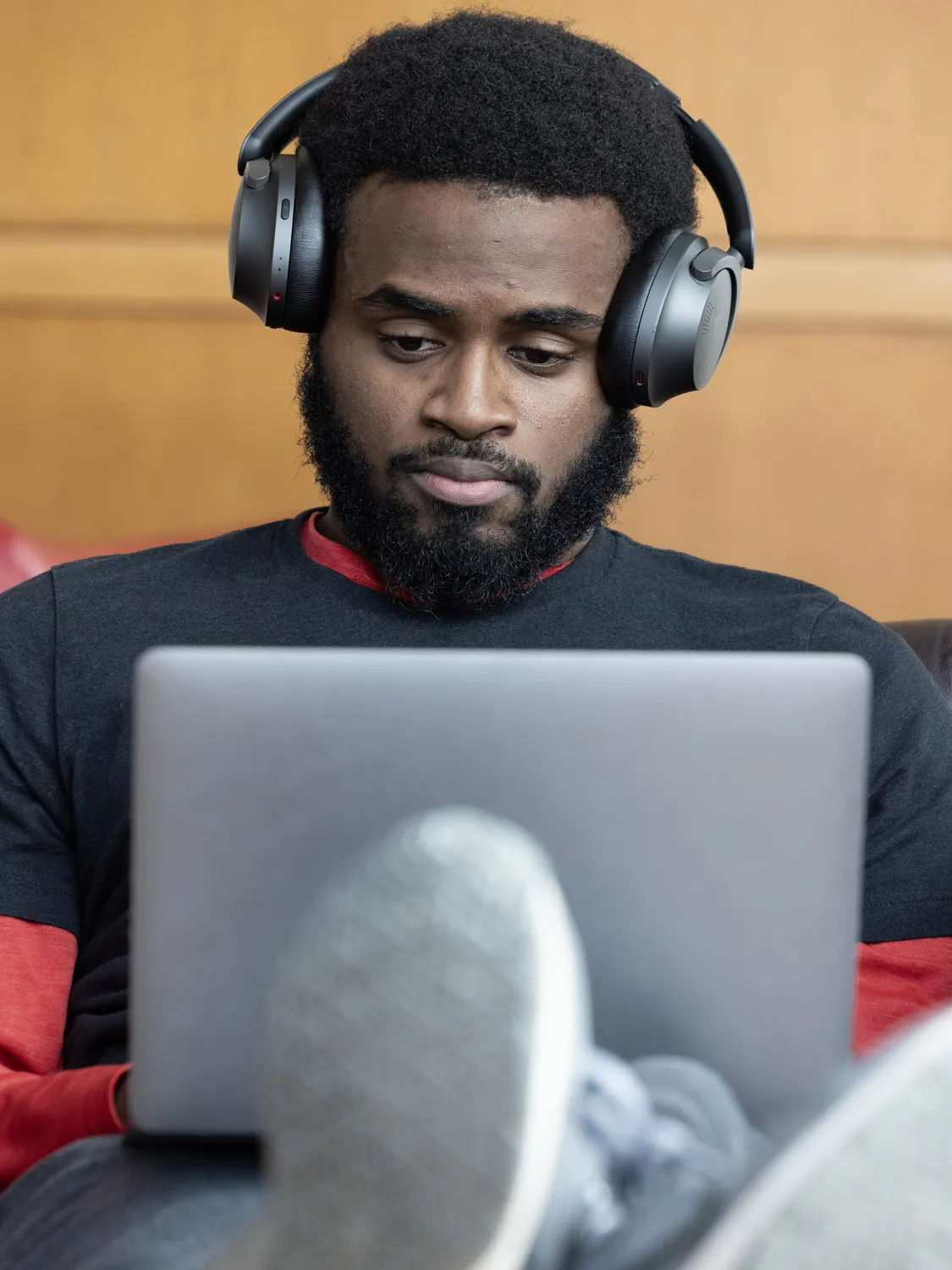 A bearded Black man, with feet propped up and his laptop on his thighs, focuses on working on his laptop. He wears big headphones, but they’re pushed above his ears, onto his temples. 
