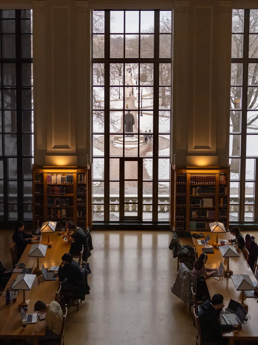 This photo, taken from high above students studying hard at long tables, shows—through tall windows—some the Oval’s iconic paths driving through the snow-covered grass sections. The light from the windows reflects off the floor and lamps here and there glow. 