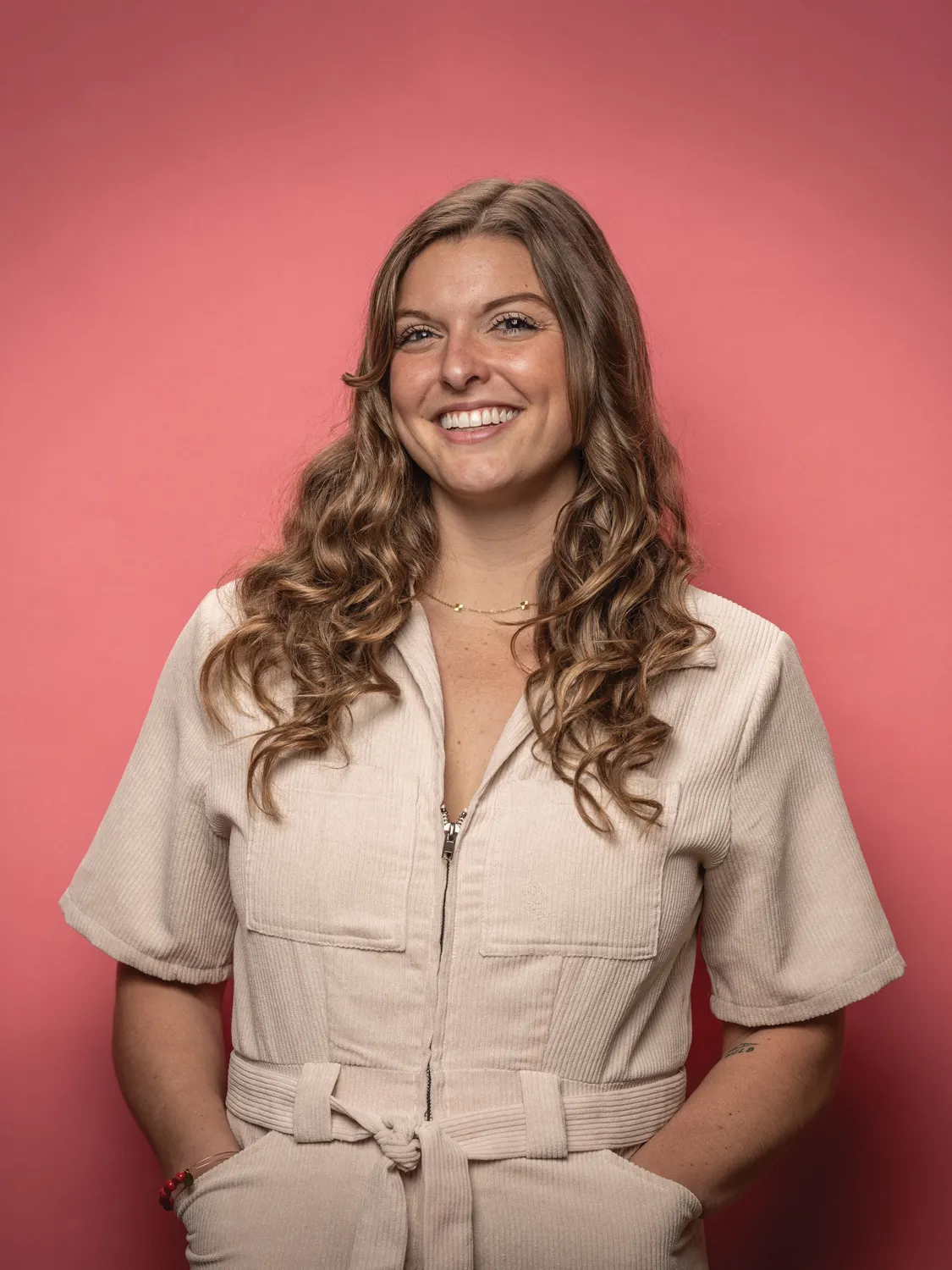 Ally Pesta smiles as she poses for a photo with her hands casually tucked in her pockets. She’s a long-haired white woman wearing a romper. 