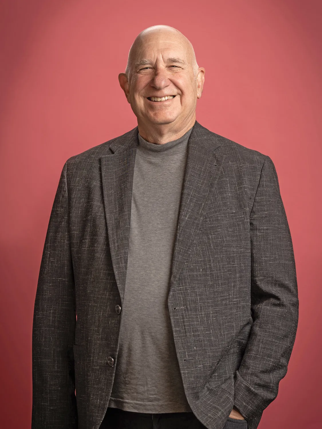 Rick Milenthal smiles as he poses for a portrait while standing up straight. He’s an older white man whose eyes squint when he grins. 