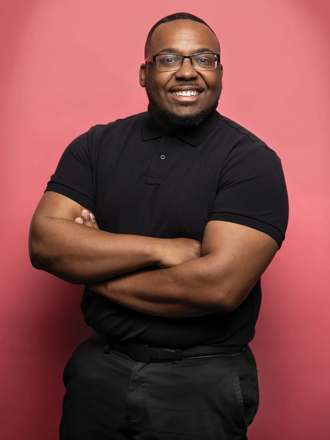 Keshawn Harper smiles as he poses for a portrait with arms folded in front of him. He’s a short-haired black man wearing glasses and a polo shirt. 