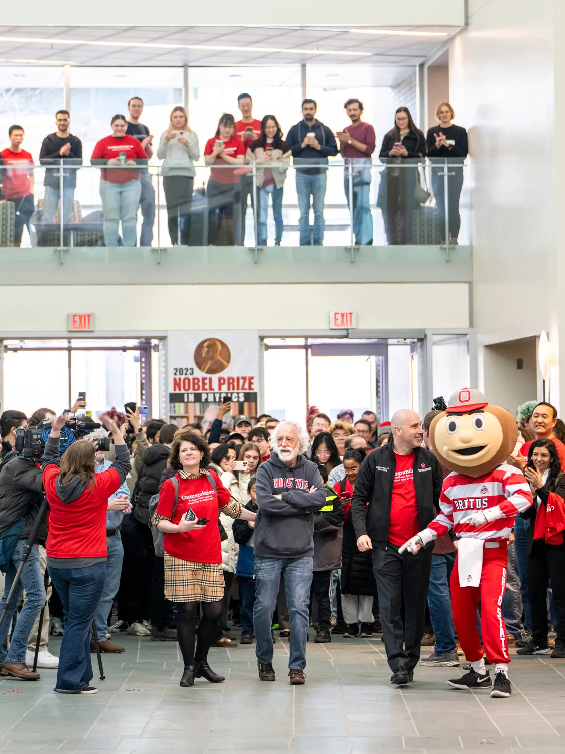 In a vertical photo showing the main floor of the Physics Research building and a balcony looking down into the lobby, crowds gather on both floors to cheer and photograph Agostini, Physics Department Chair Michael Poirier and Brutus Buckeye, a person dressed in Ohio State colors and wearing a giant, smiling, buckeye nut-shaped head. 