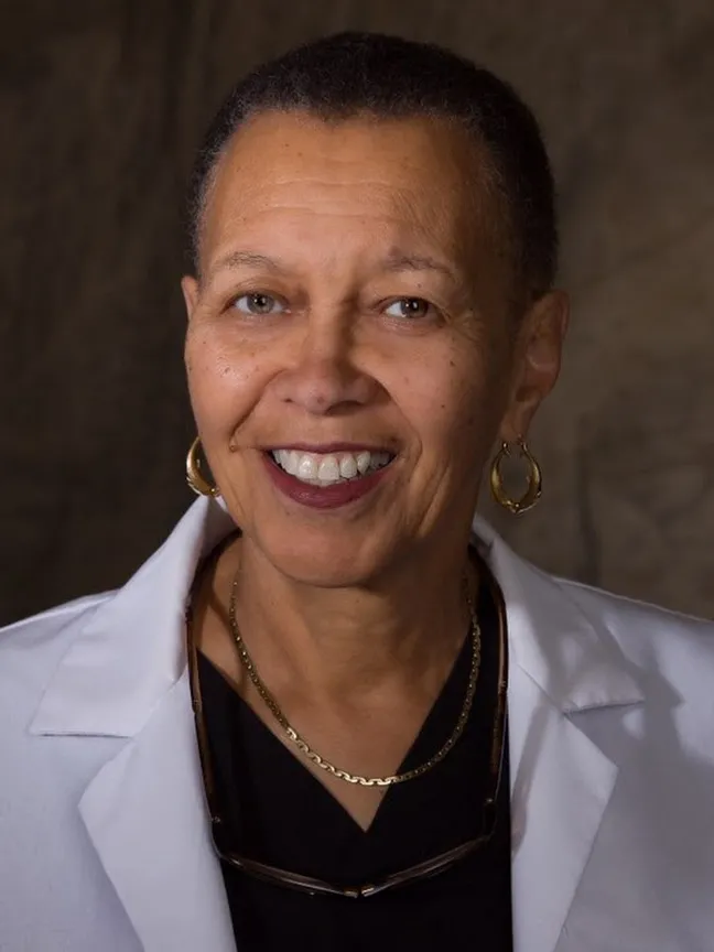 In a portrait showing her head and shoulders, Romaine Bayless smiles while wearing her lab coat, gold hoops earrings and a pair of gold necklaces. A black woman with close cropped hair, a pretty and wide smile, and eyes that crinkle as the corners, her expression says she is friendly and smart.