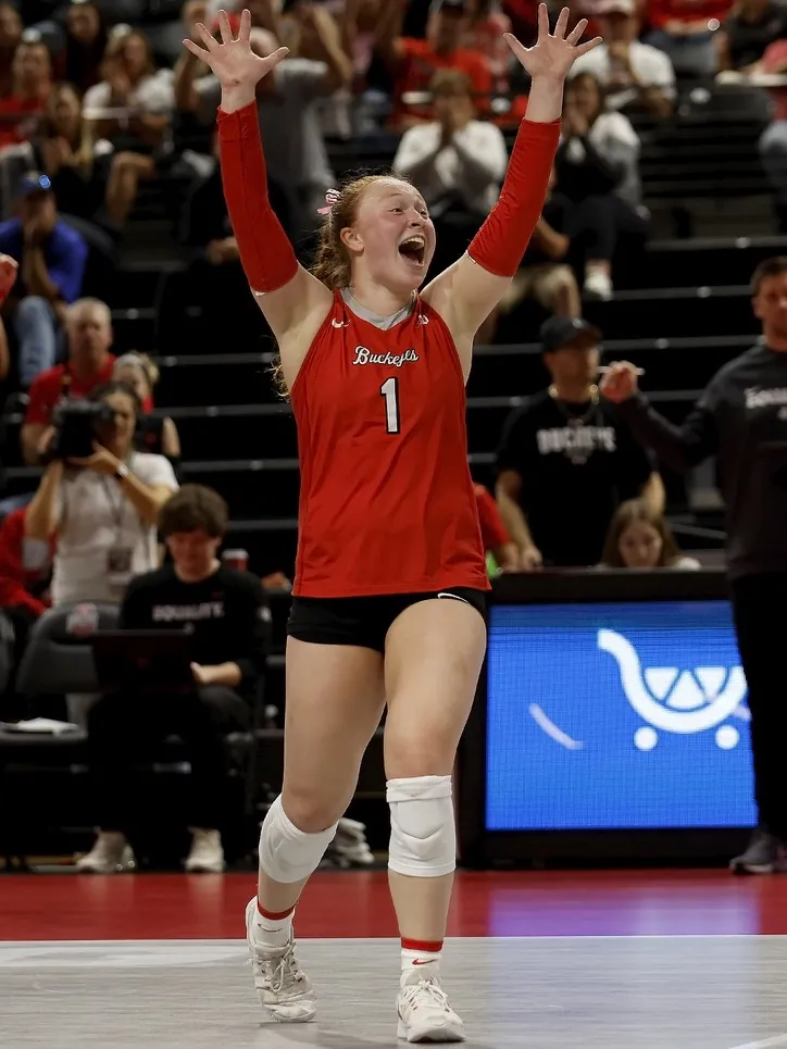 Sarah Morbitzer, a young white woman with her red hair pulled back in a braid, cheers a good play while on the volleyball court. She wears an Ohio State volleyball uniform, with sleeveless shirt, short black shorts, kneepads and arm wraps. The stands and fans are in the background.