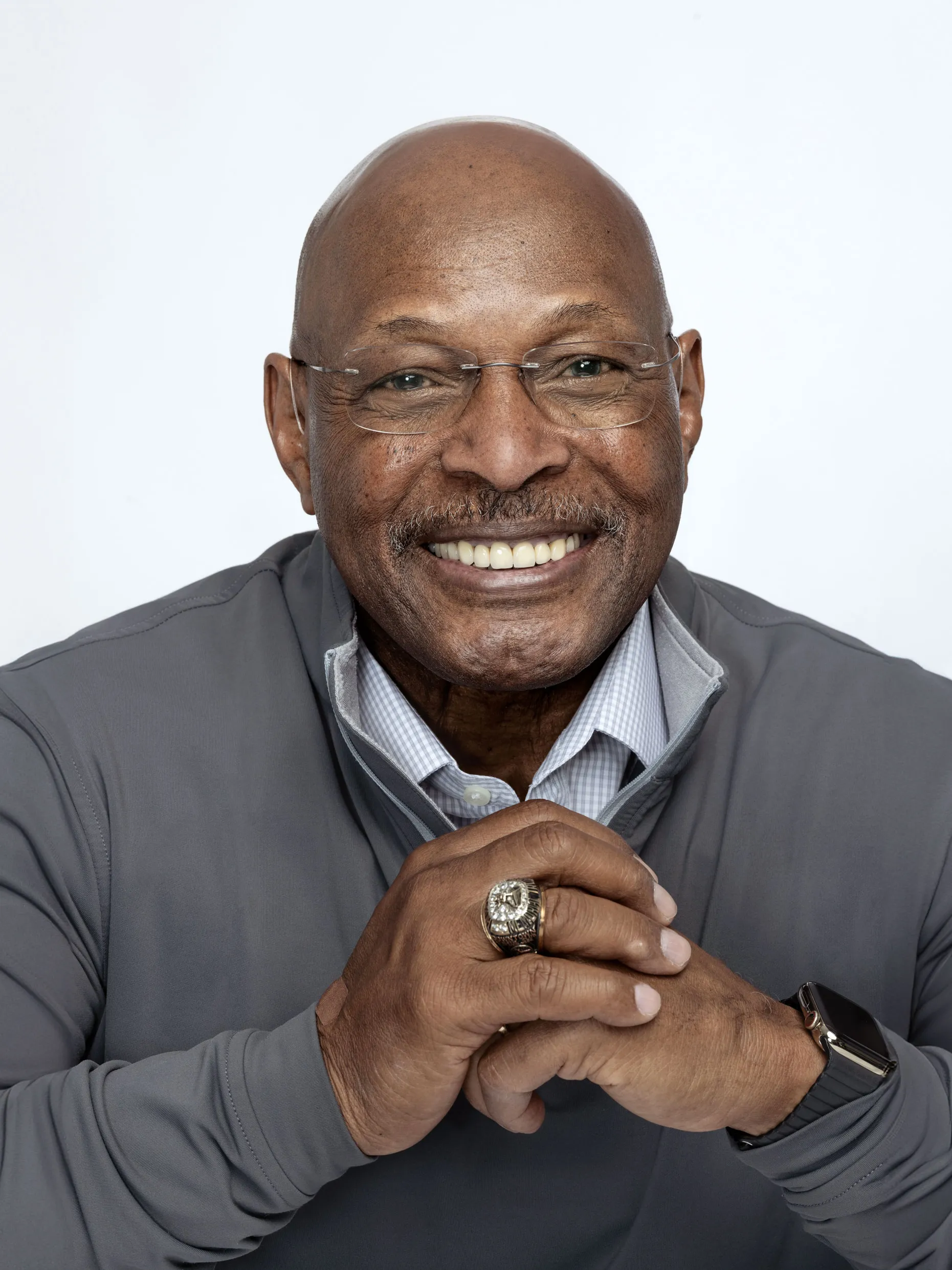 Archie Griffin, a 70-year-old Black man, smiles big for the camera with his hands clasped in front of his chest. The pose shows off his Heisman ring. The famous Buckeye looks happy and friendly. 