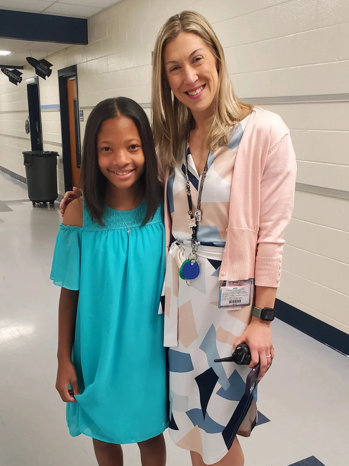 A student and teacher pose for this photo in a school hallway. They’re smiling and each has an arms wrapped around the other—their closeness speaks to their good relationship. The teacher is a tall white woman with long blond hair and a stylish dress. The student is a black girl wearing a pretty dress. 