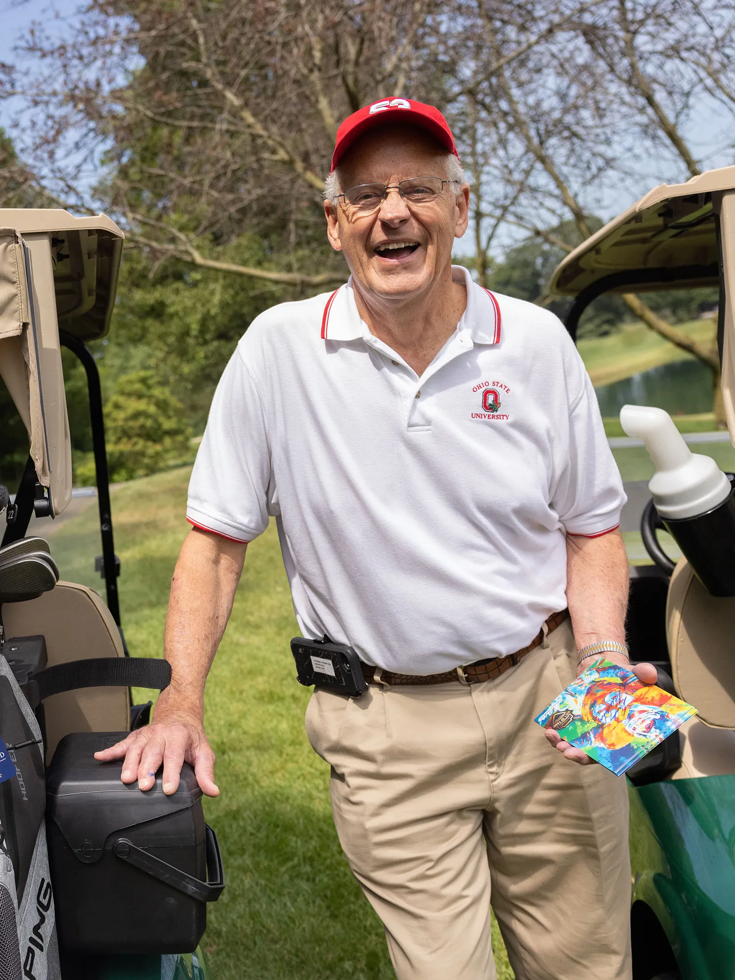 Randy Gradishar, an older white man wearing golfing clothes and a ballcap, slightly leans on a golf cart as he laughs during a conversation with someone not shown in the photo.  