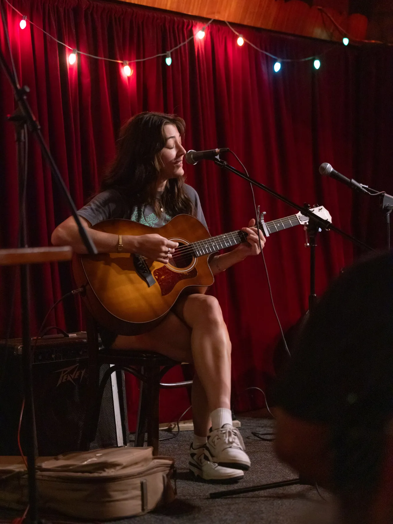 While sitting on a stool, Tovah Kaiser sings with her eyes closed as she strums an acoustic guitar plugged into an amp that is outside of the photo frame. A mic is set up at mouth height and behind her is a red curtain with a string of large Christmas lights. She’s in sneakers and a T-shirt. 